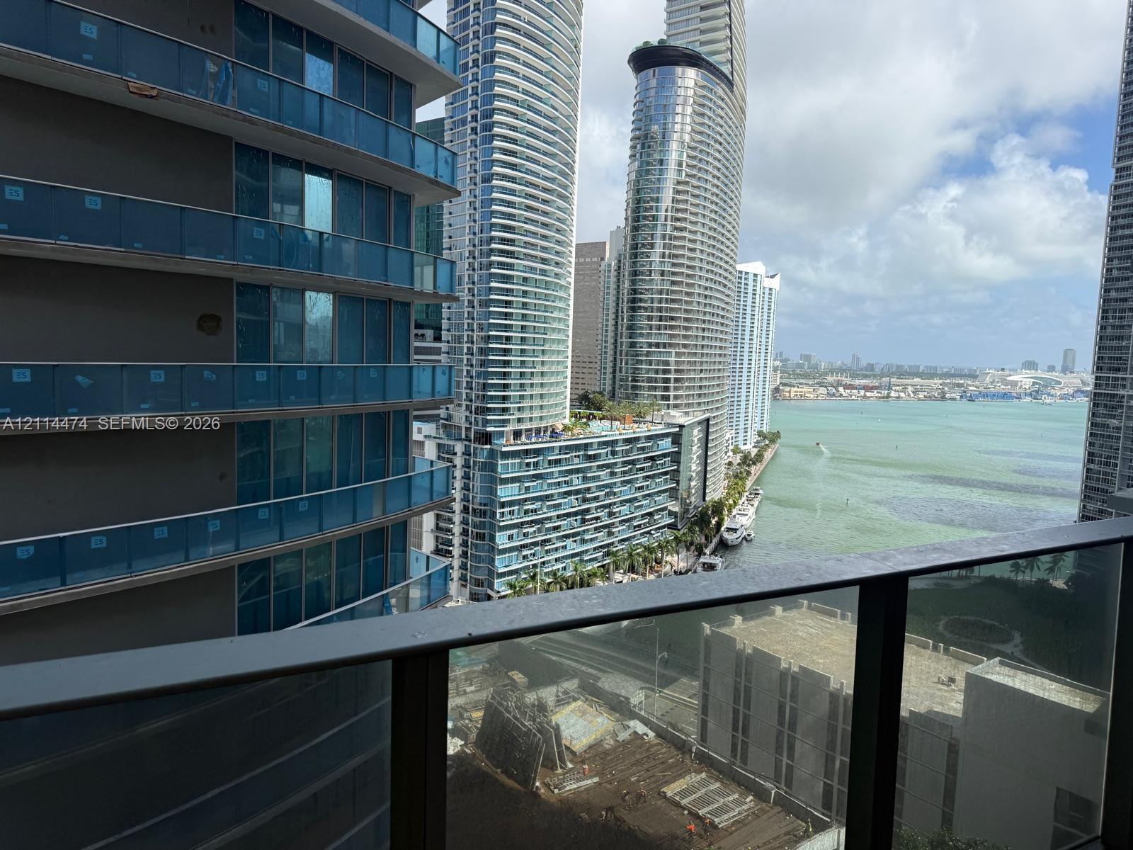 Balcony featuring metal railings with glass panels overlooking a bay, high-rise urban structures, and waterfront scenery