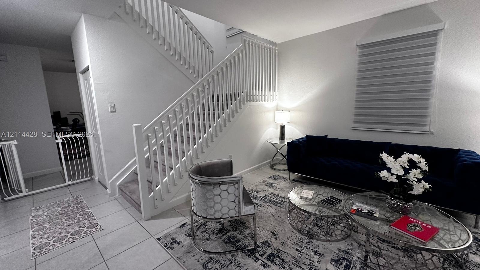 Open-concept living space featuring neutral ceramic tile flooring, a staircase with custom white vertical balusters, and tiered window treatments