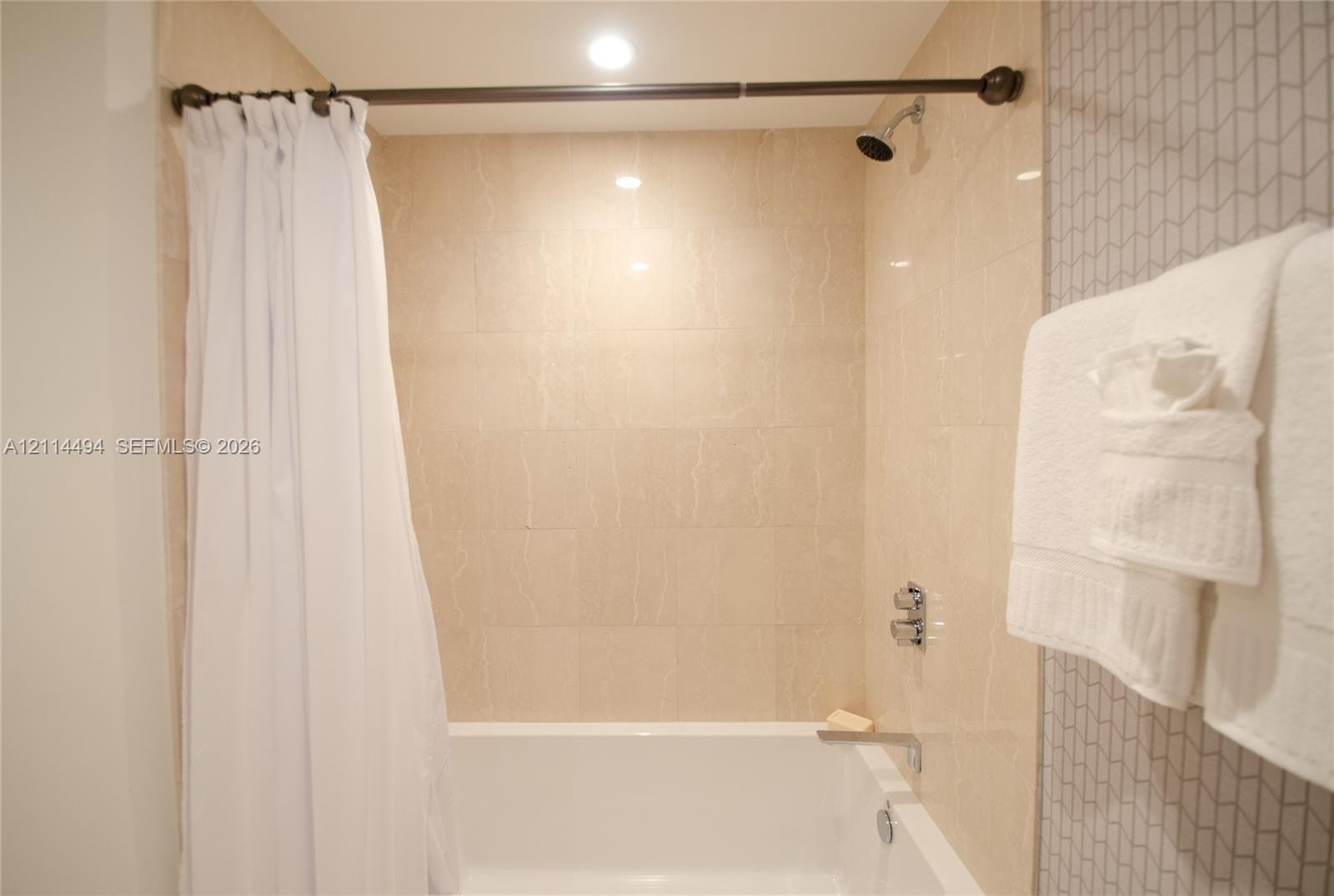 Shower and bathtub combination featuring large-format beige wall tiles, chevron-patterned accent tiling, a chrome wall-mounted shower head, and a chro