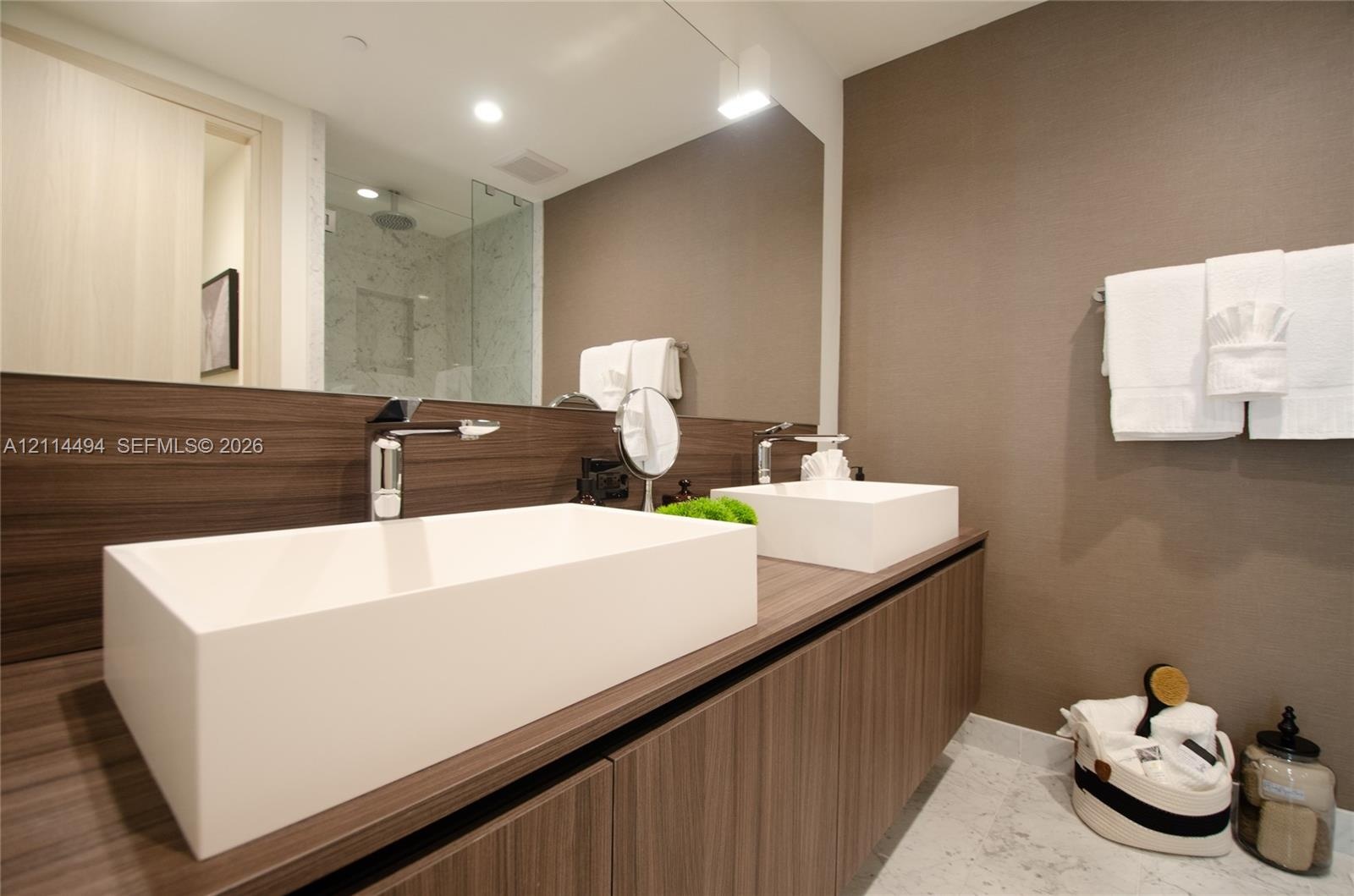 Floating wood-finish vanity with dual rectangular vessel sinks, polished chrome faucets, large frameless mirror, and walk-in shower with stone-finish 