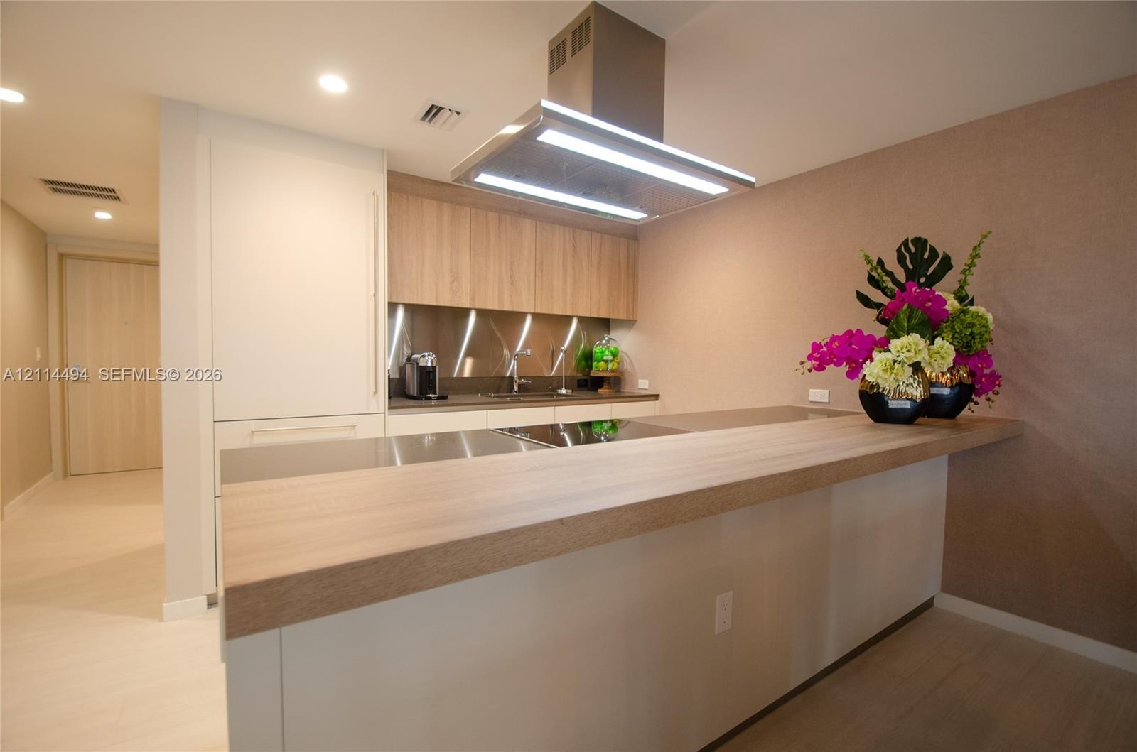 Modern kitchen featuring wood-finish cabinetry, sleek stainless steel backsplash, integrated cooktop, extended breakfast bar, and a stainless steel is
