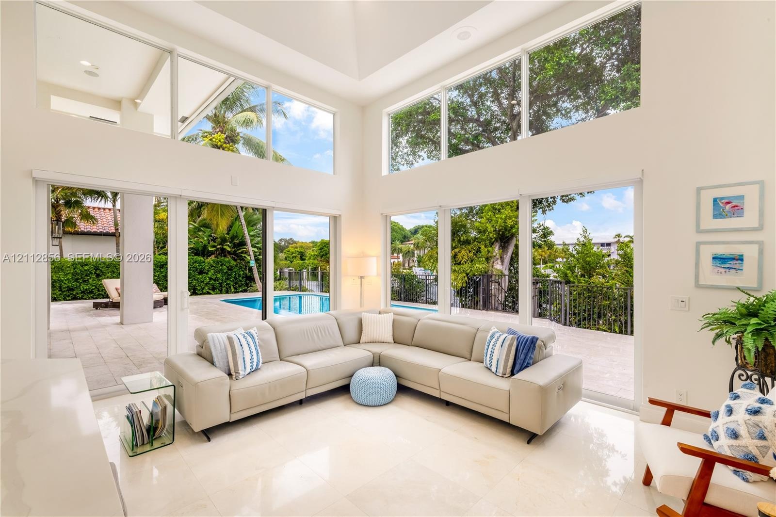 Double-height ceilings, floor-to-ceiling windows, polished stone flooring, and multiple sliding glass doors opening to a paved patio with a swimming p
