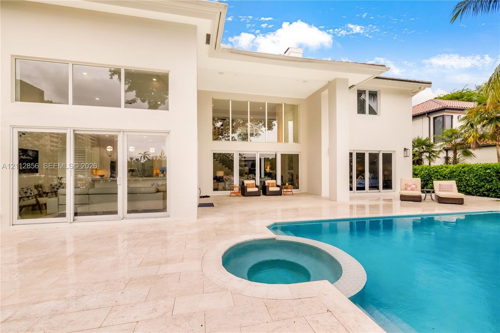 Exterior elevation featuring a stucco facade, expansive glass walls with multi-panel sliding doors, a stone-paved patio, and a swimming pool with a bu