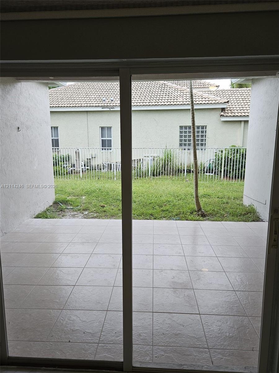 Covered patio with neutral-toned square tile flooring, enclosed exterior space with a white metal fence, and grass lawn