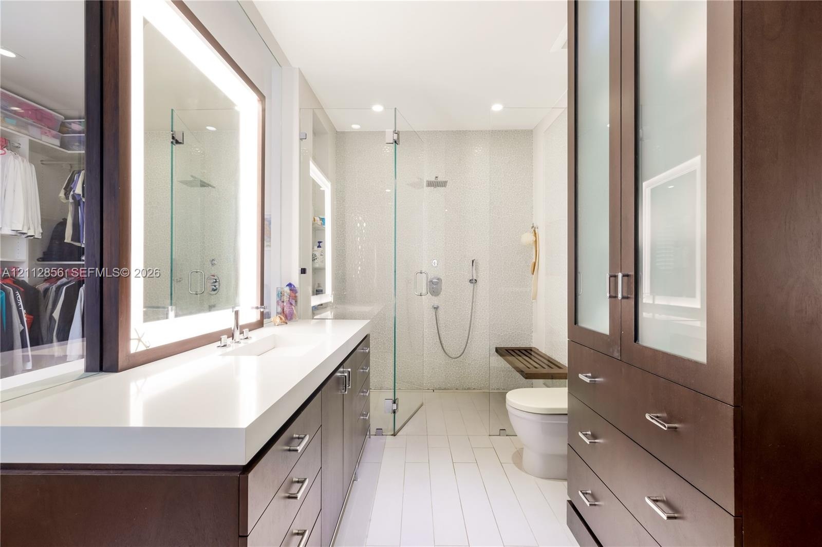 Custom dark-wood cabinetry, white composite countertops, integrated LED-backlit mirror, glass-enclosed walk-in shower with pebble-tiled wall, and buil