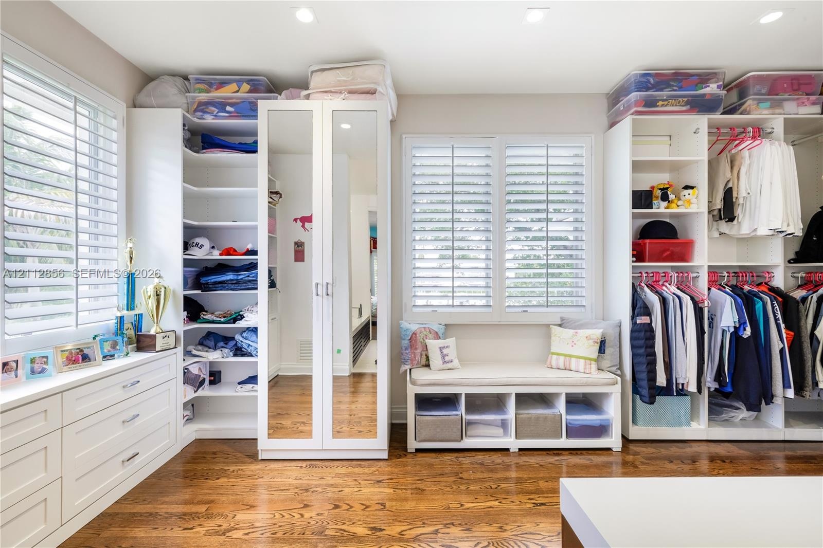 Custom closet featuring white built-in cabinetry, floor-to-ceiling mirrored doors, integrated window seating, plantation shutters, and wood-finish flo