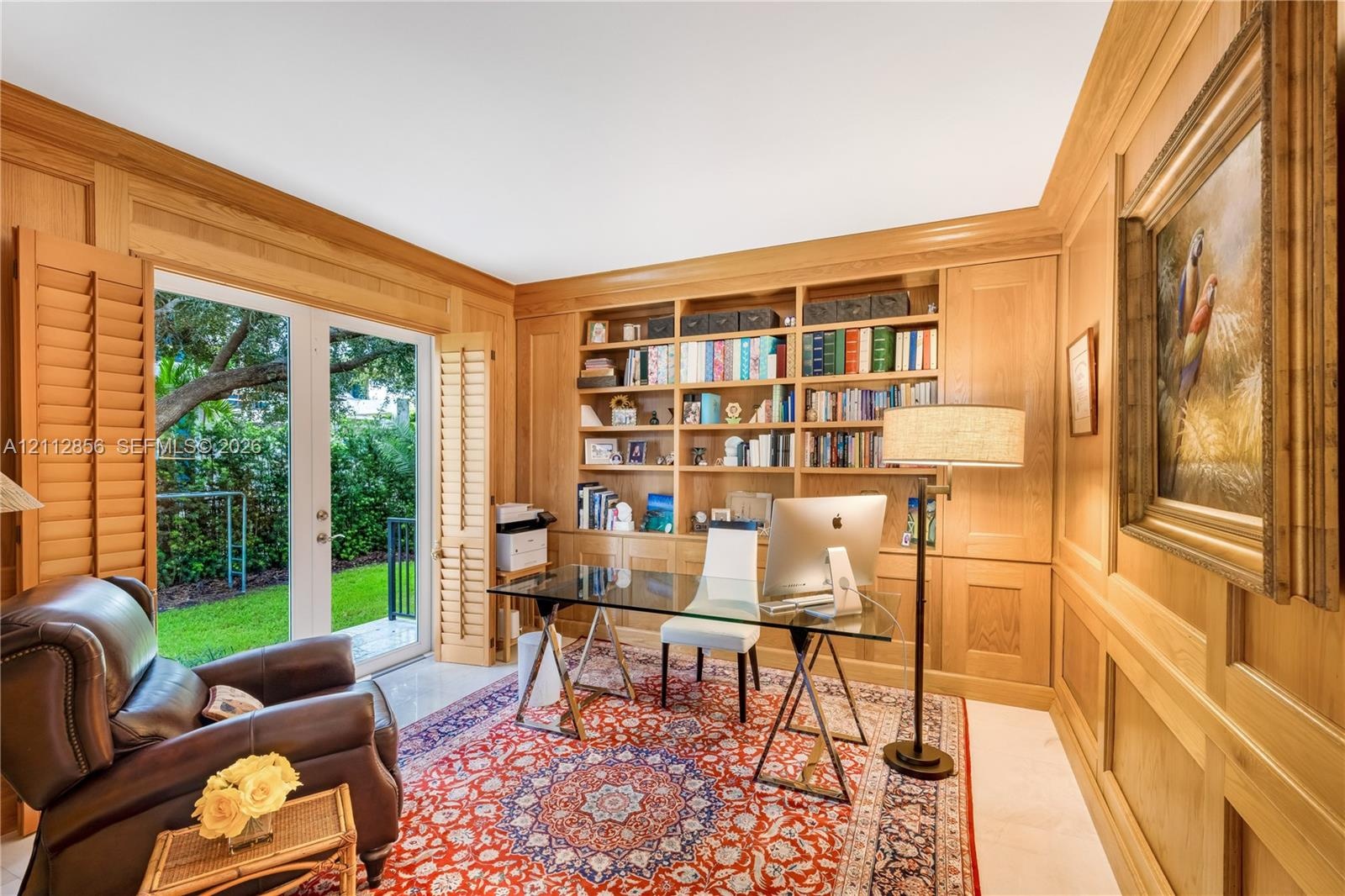 Wood-paneled room featuring custom floor-to-ceiling built-in bookshelves, light-finish tile flooring, and glass French doors with integrated louvered 