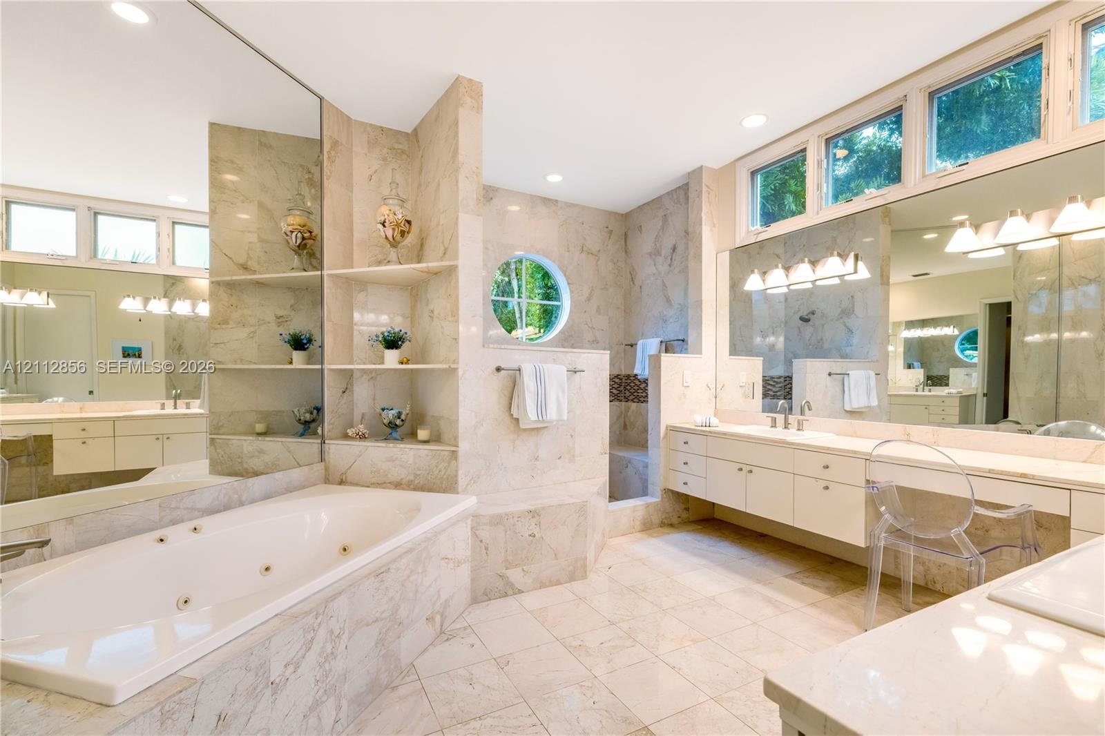Spacious bathroom featuring stone-tiled surfaces, a built-in jetted tub, a circular accent window, dual vanities with integrated cabinetry, and extens
