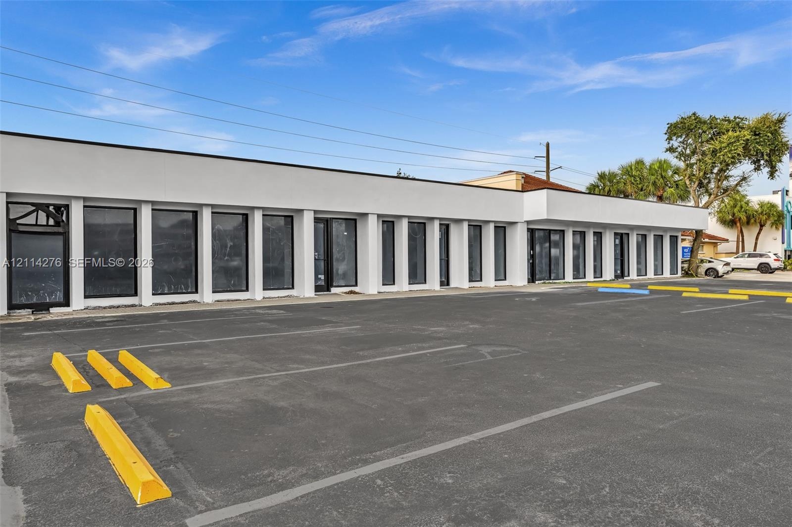 Commercial exterior featuring a flat roofline, floor-to-ceiling glass windows, a white facade, and an asphalt parking lot with designated wheel stops