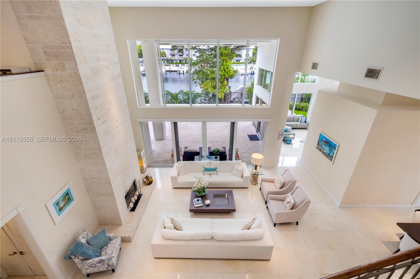 Double-height living area with natural stone fireplace surround, floor-to-ceiling windows, light-toned tile flooring, and direct access to an outdoor 