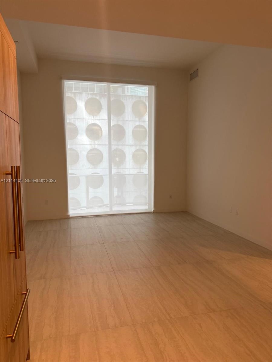 Large-format tile flooring, floor-to-ceiling window with privacy screen, and built-in wood-finish cabinetry featuring vertical bar pulls