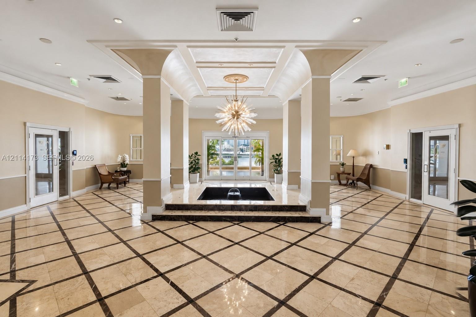 Grand lobby featuring polished stone flooring with geometric tile inlay, a central tiered water feature, soaring architectural pillars, recessed tray 
