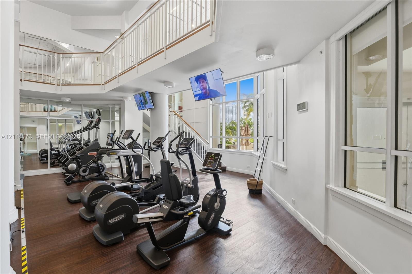 Two-story fitness space featuring wood-finish flooring, floor-to-ceiling windows, an elevated mezzanine railing, and expansive mirrors