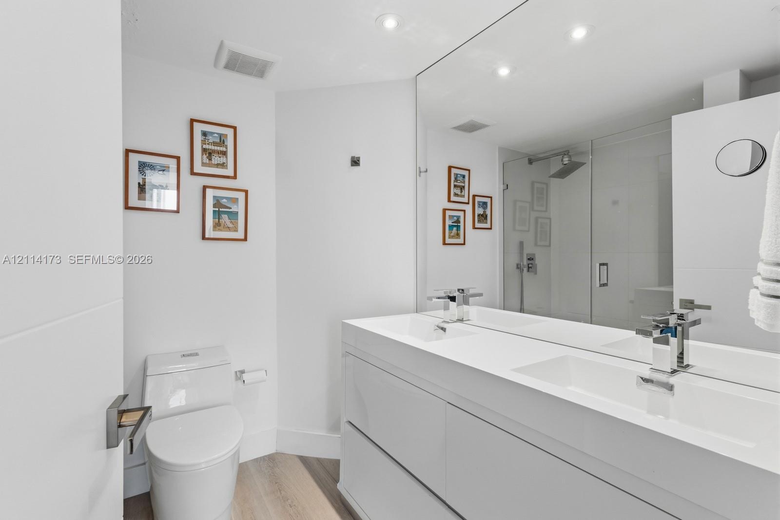 Dual-vanity cabinet with integrated rectangular sinks, expansive wall-to-wall mirror, glass-enclosed shower, and wood-finish flooring