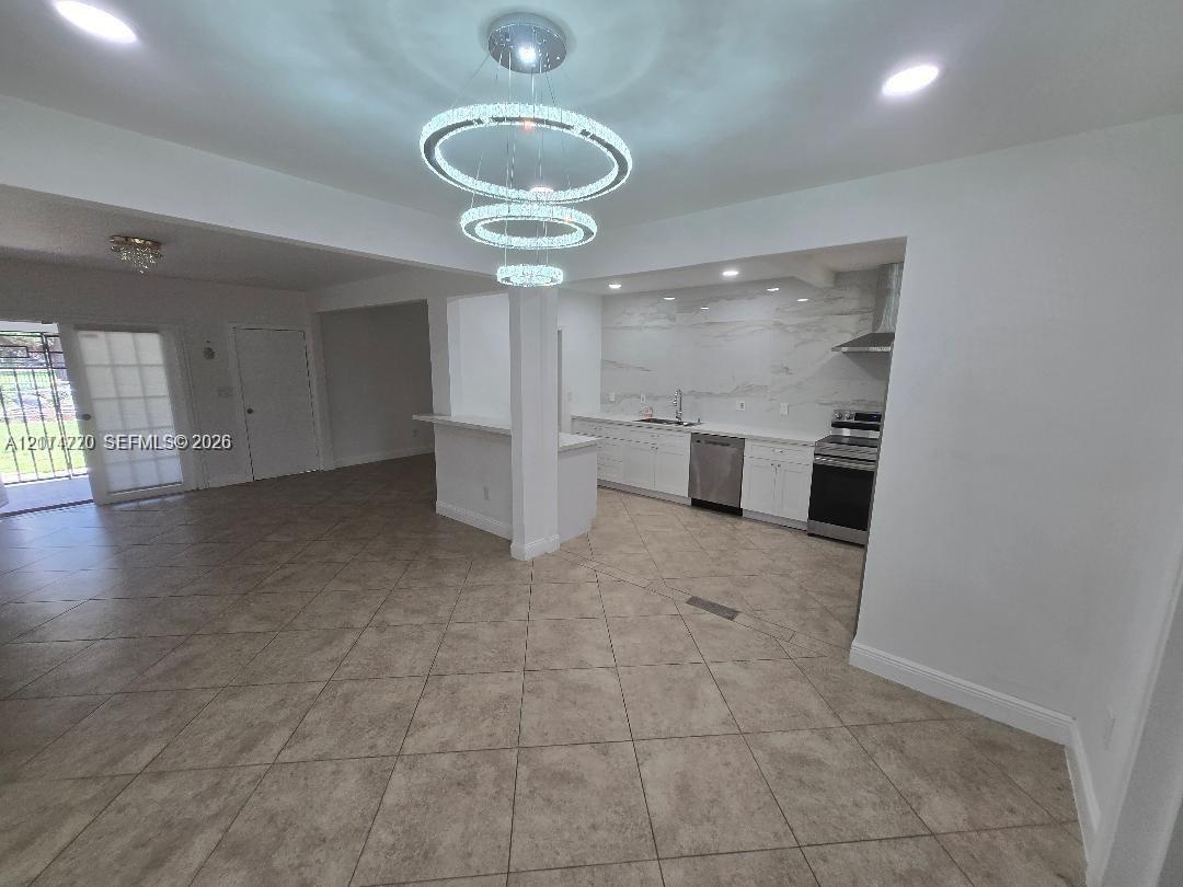 Open-concept kitchen featuring white cabinetry, stone-patterned slab backsplash, stainless steel appliances, tiered crystal chandelier, and tile floor
