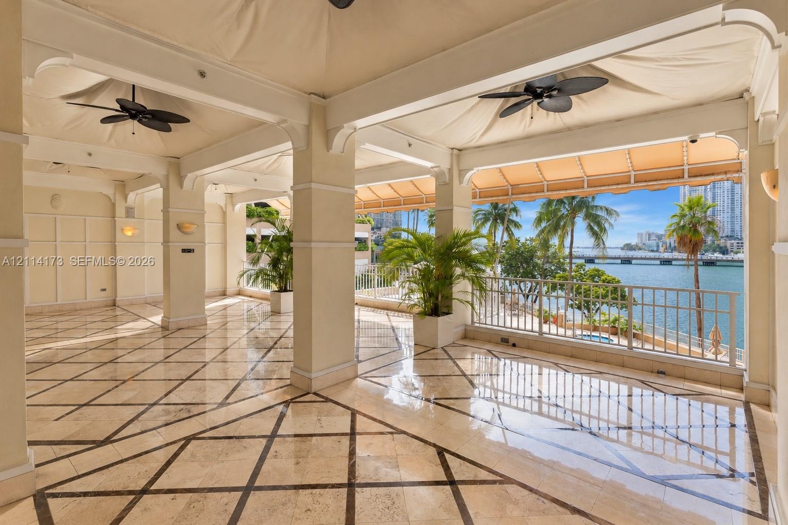 Covered terrace featuring stone tile flooring with decorative inlay, structural columns, ceiling-mounted fans, and metal railings overlooking the wate