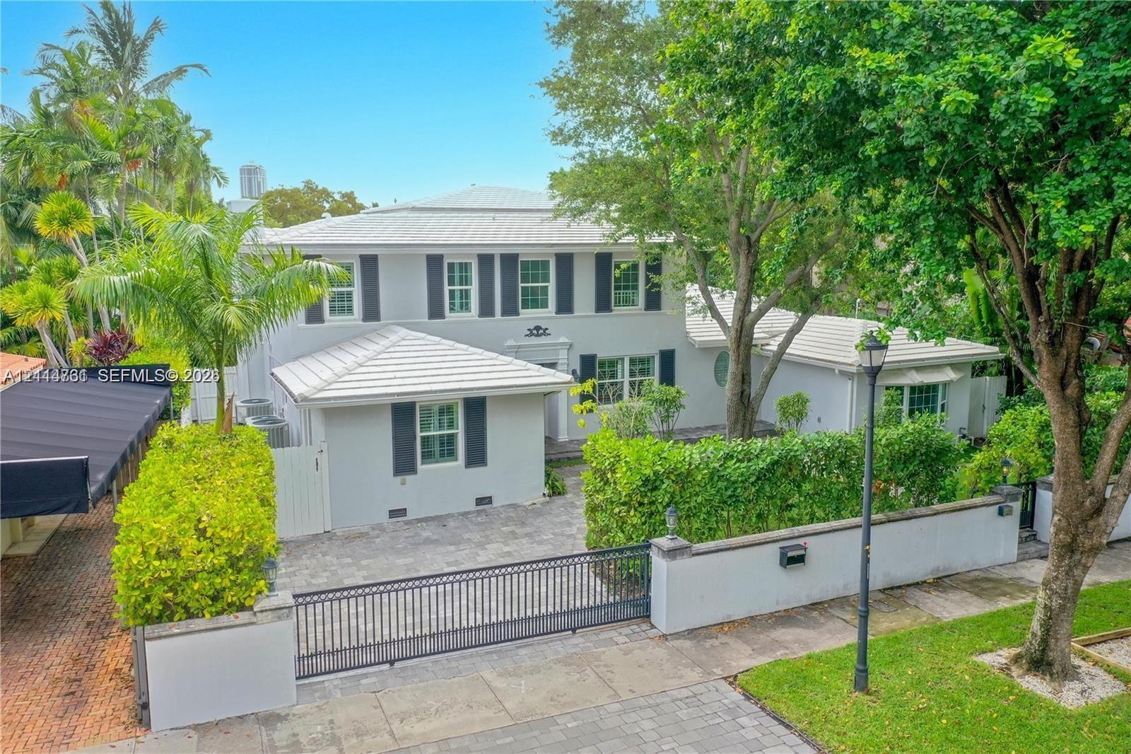 Two-story exterior featuring a light-toned stucco façade, metal roof, shuttered windows, paver driveway, and a gated perimeter wall