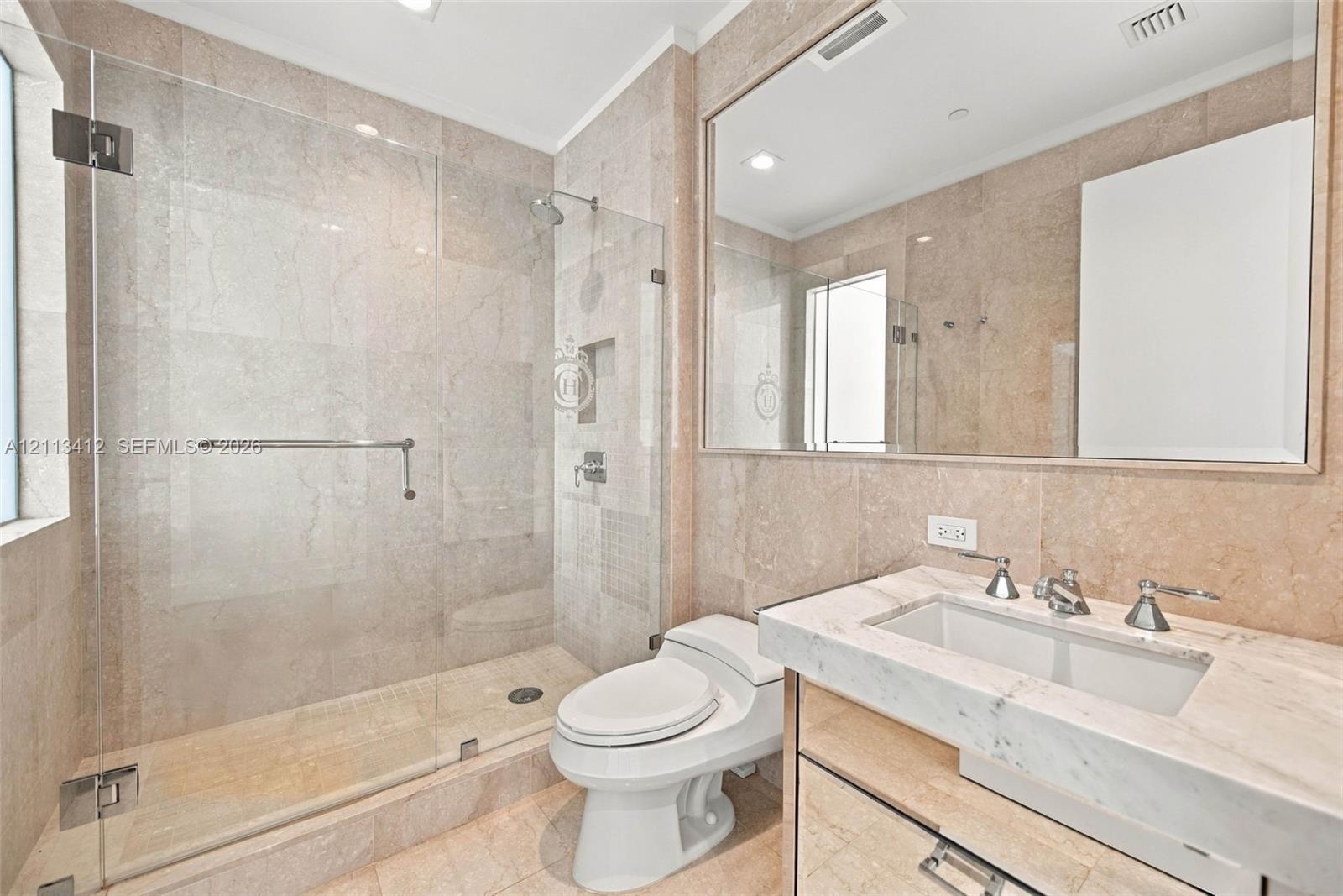 Full-wall stone tiling, glass-enclosed walk-in shower with built-in recessed niche, vanity with integrated basin, polished stone countertop, and large
