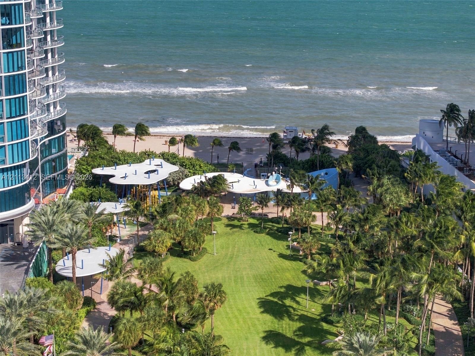 Expansive coastal grounds featuring manicured green space, mature palm trees, paved walking paths, and organic-shaped shade structures