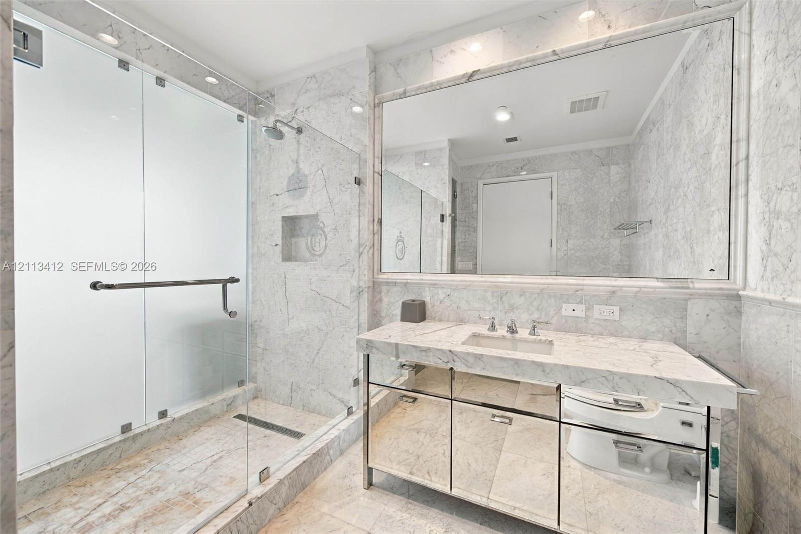 Full-height stone wall surfacing, frameless glass shower enclosure, vanity with integrated sink, mirrored cabinet base, and wall-to-wall vanity mirror