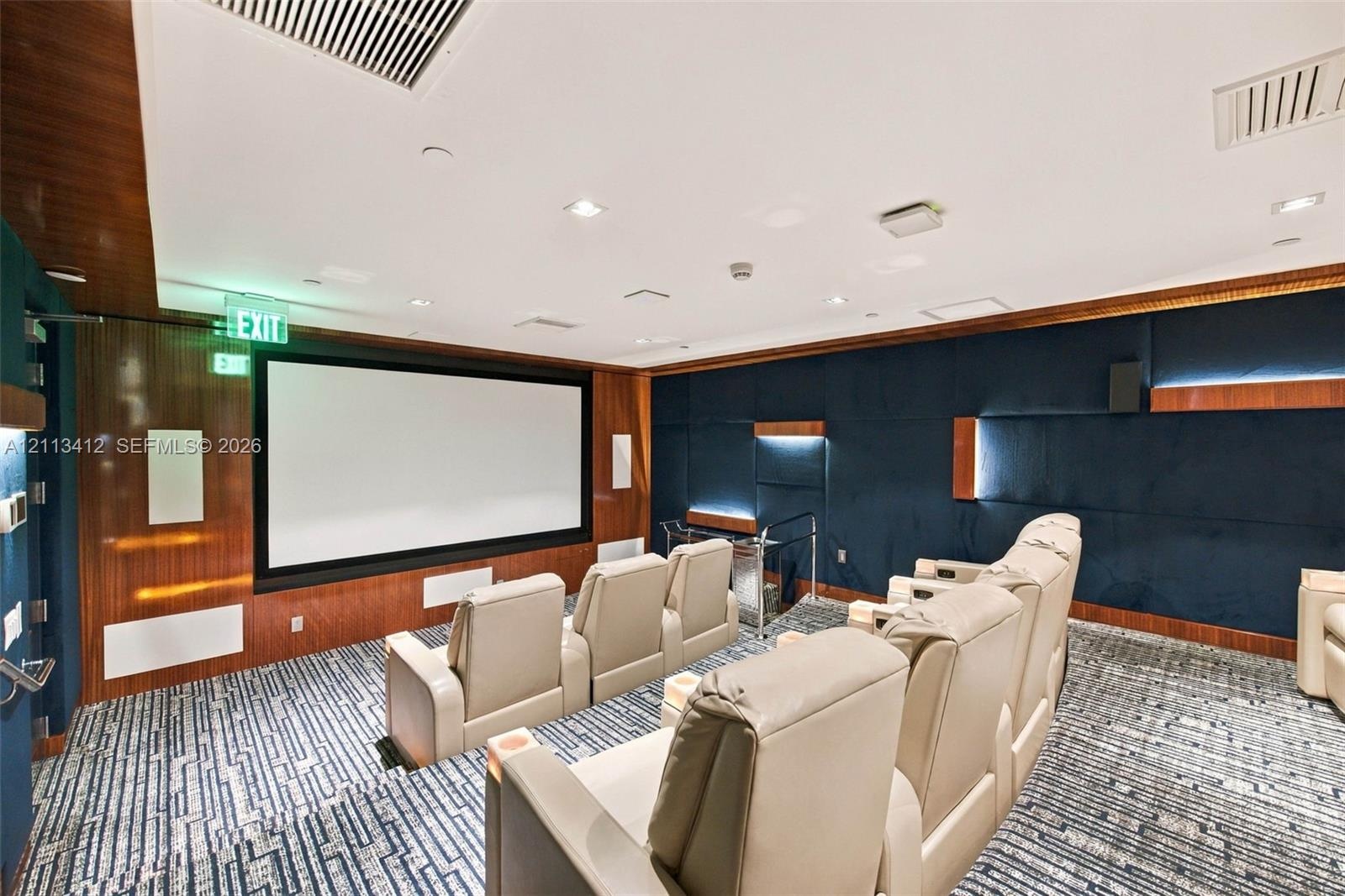 Media room featuring tiered flooring, dark blue upholstered wall panels, integrated wood-finish wall accents, recessed lighting, and a built-in projec