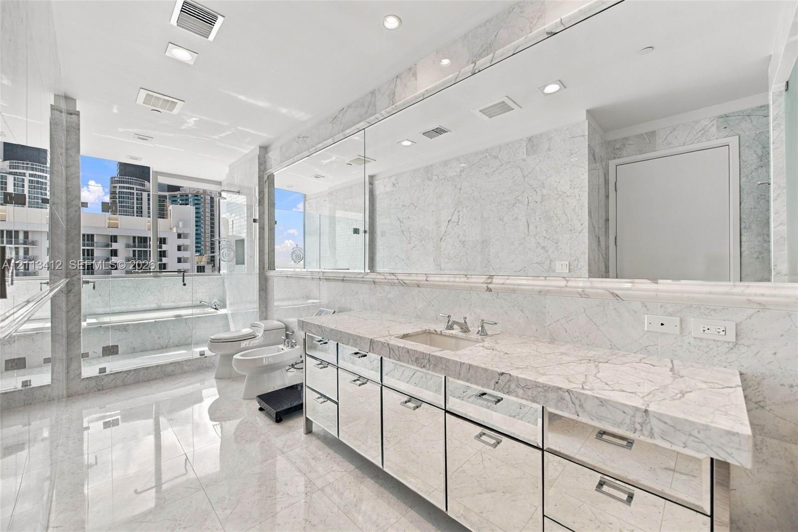 Full-length stone vanity with dual undermount sinks, expansive wall-to-wall mirror, floor-to-ceiling natural stone wall and floor tile, glass-enclosed