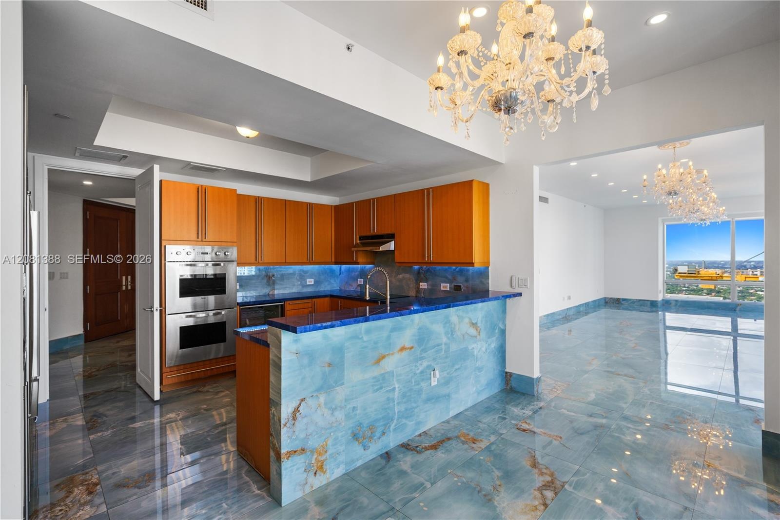 Kitchen featuring natural stone flooring, matching stone-clad peninsula, built-in double wall oven, wood-finish cabinetry, and decorative crystal chan