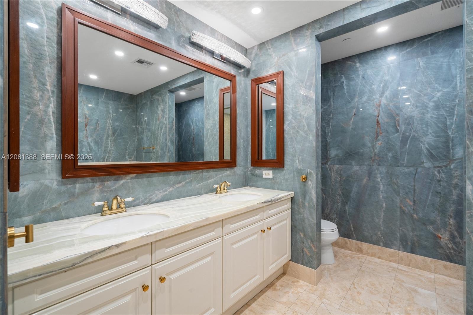 Double vanity with white cabinetry, stone-finish countertops, brass-toned fixtures, wood-framed mirrors, and floor-to-ceiling stone wall cladding