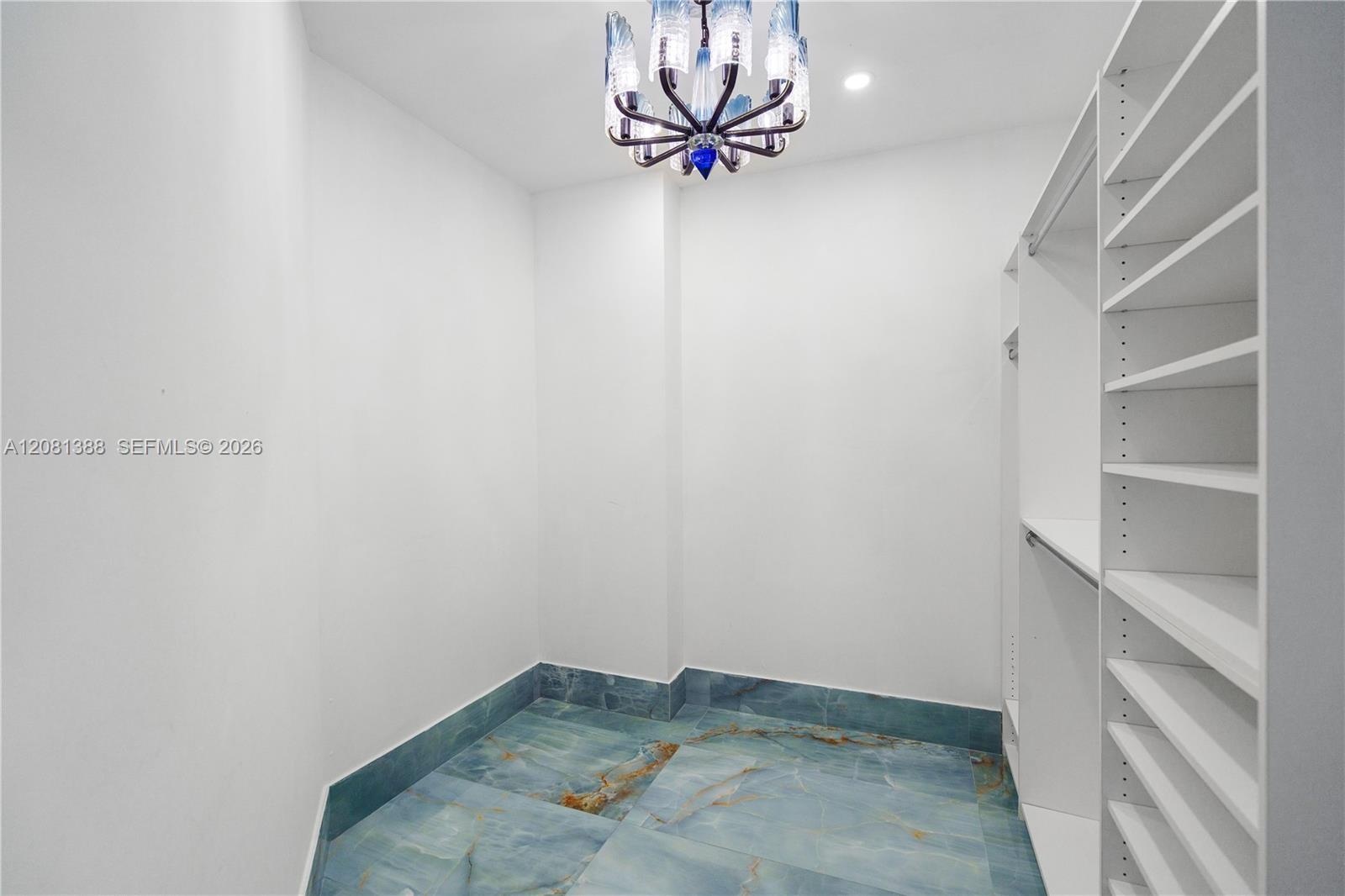 Walk-in storage space featuring built-in white shelving, recessed lighting, a decorative chandelier, and large-format polished stone flooring