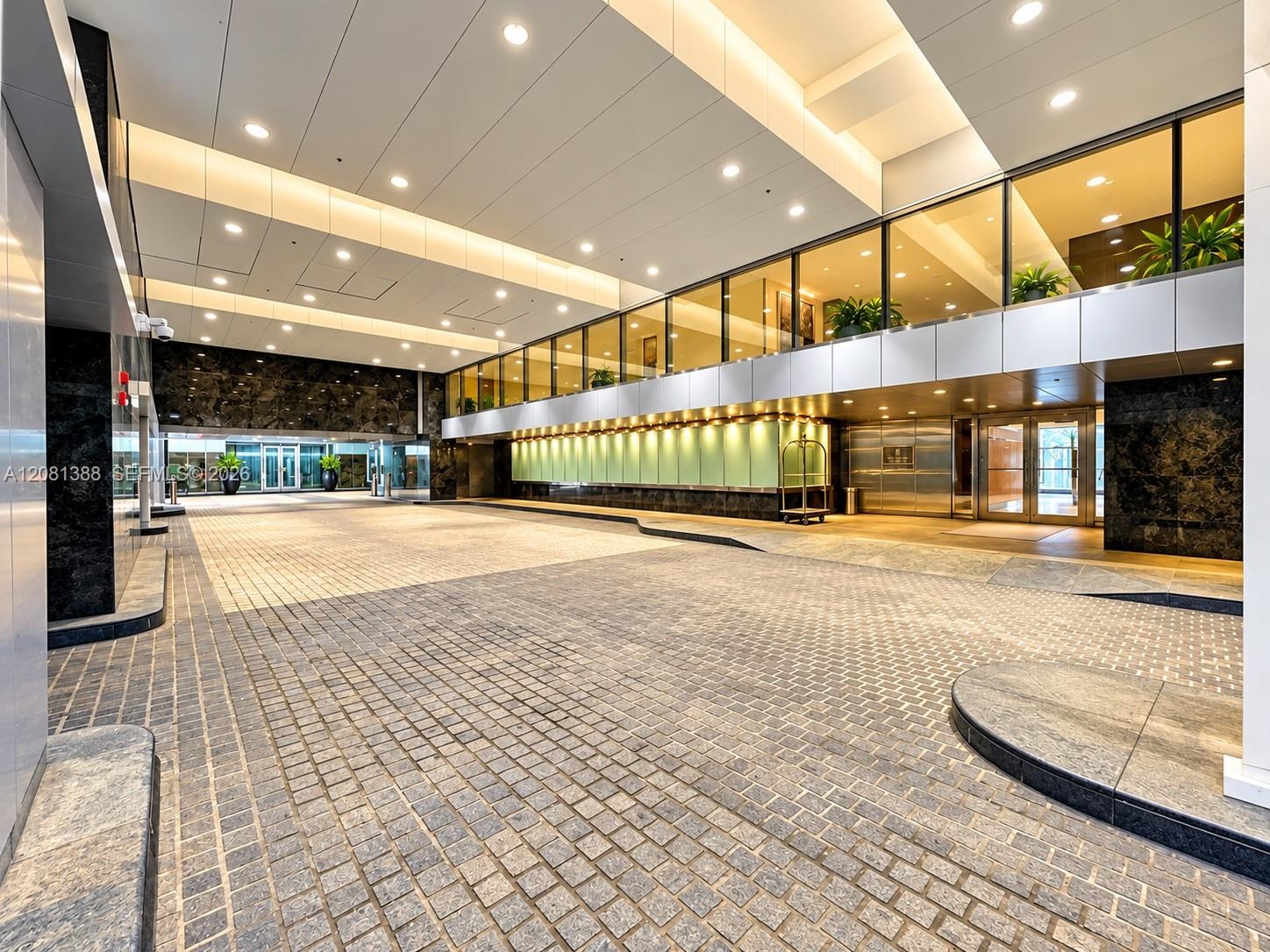 Covered motor court entrance featuring stone paver flooring, dark polished stone wall accents, extensive recessed lighting, and a glass-enclosed upper