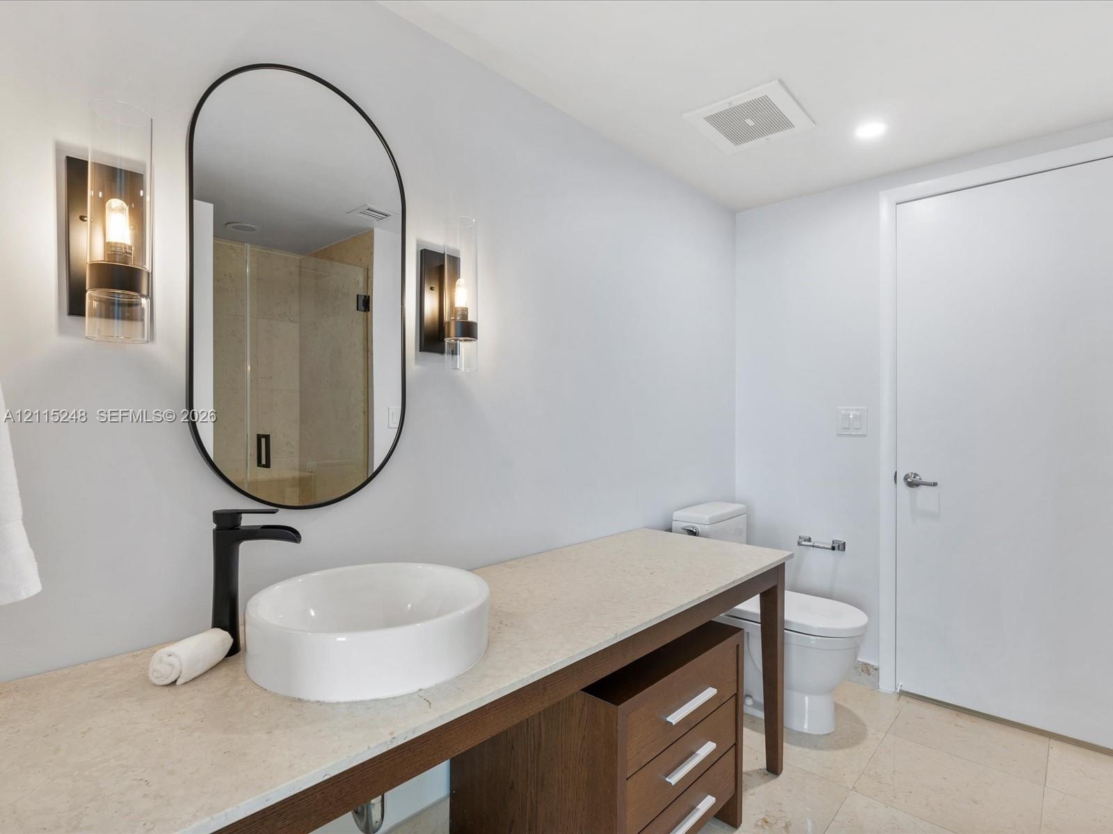 Vanity featuring a stone-finish countertop, round vessel sink, matte black faucet, oval wall-mounted mirror, and tiered wood cabinetry