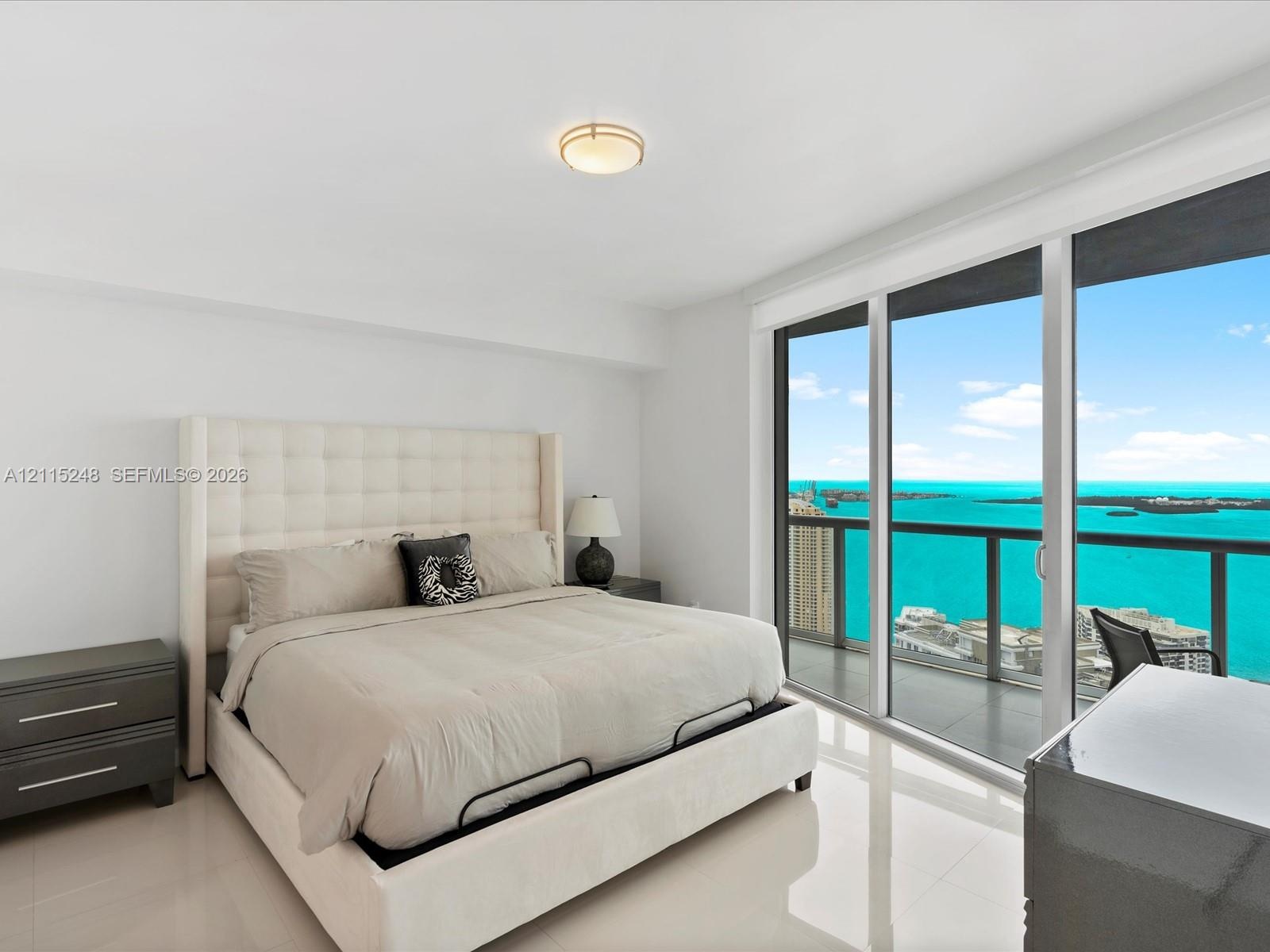 Floor-to-ceiling glass sliding doors, private balcony with metal railing, neutral tile flooring, flush-mount circular lighting fixture, and panoramic 