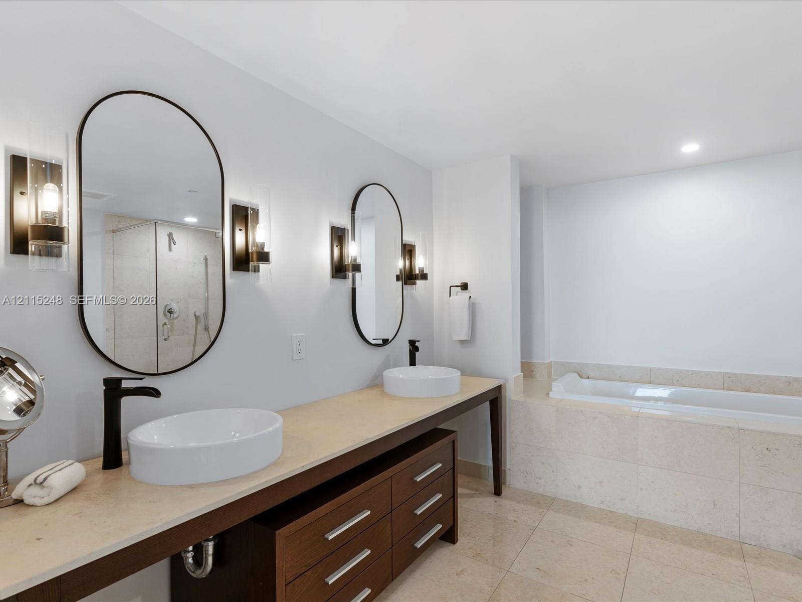 Dual vanity featuring stone-surfaced countertops, vessel sinks with matte black faucets, wood-finish cabinetry, and oval mirrors with integrated wall-