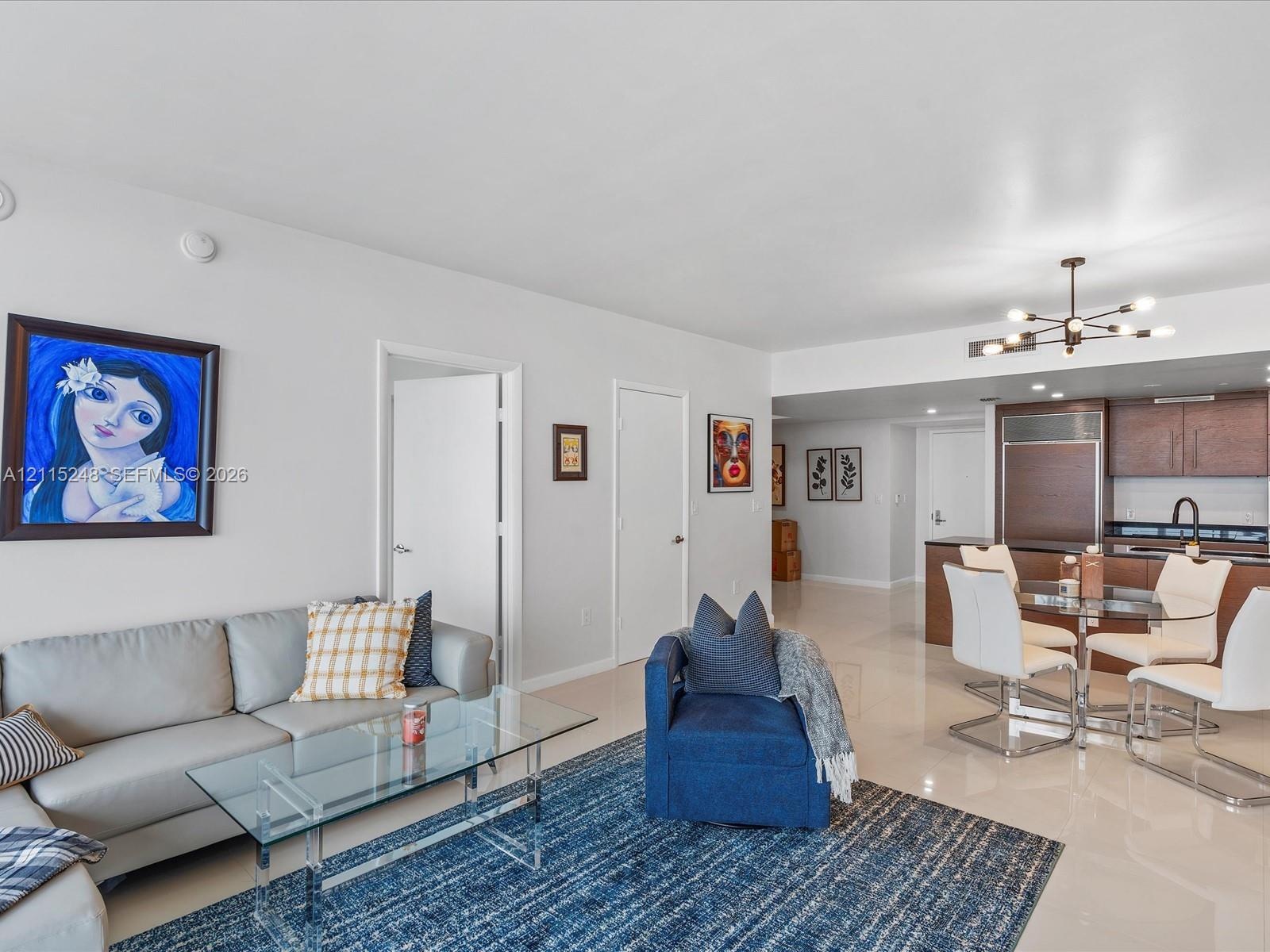 Open-concept interior featuring light-toned tile flooring, recessed ceiling lighting, a modern multi-arm chandelier, and a built-in cabinetry kitchen 