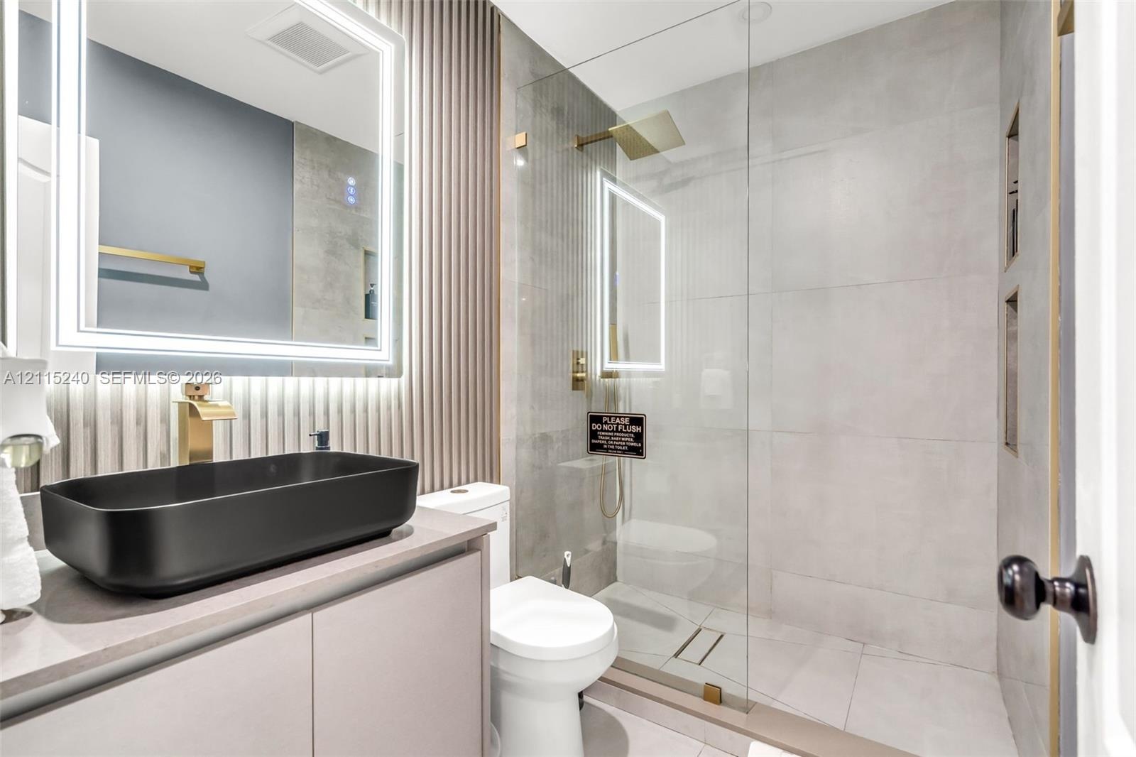 Bathroom featuring a matte black vessel sink, gold-toned faucet hardware, wall-mounted LED-backlit mirror, vertical slat accent wall, and a walk-in sh