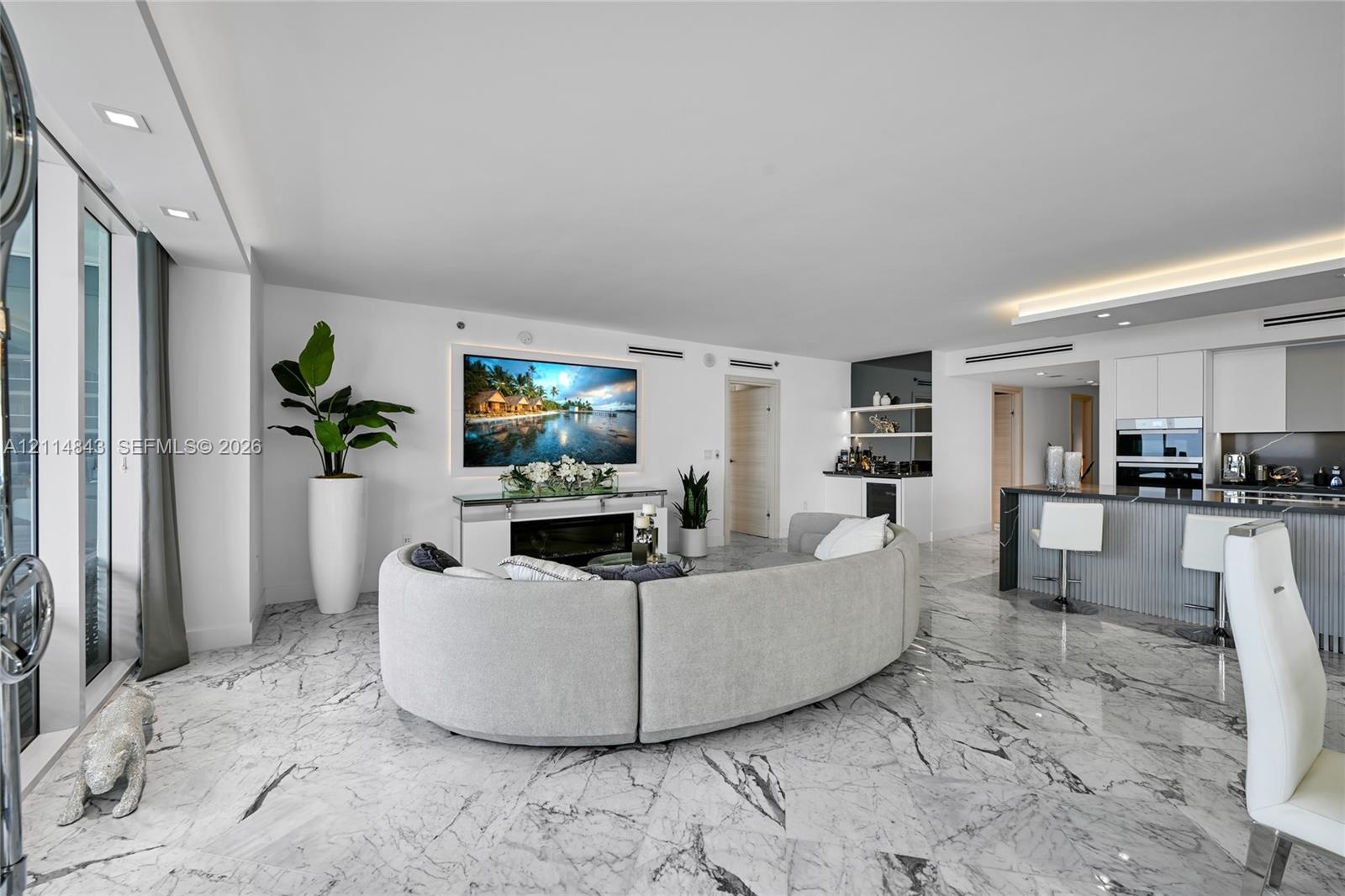 Open-concept living space featuring white stone-finish flooring, recessed lighting, built-in electric fireplace, recessed wet bar with shelving, and k