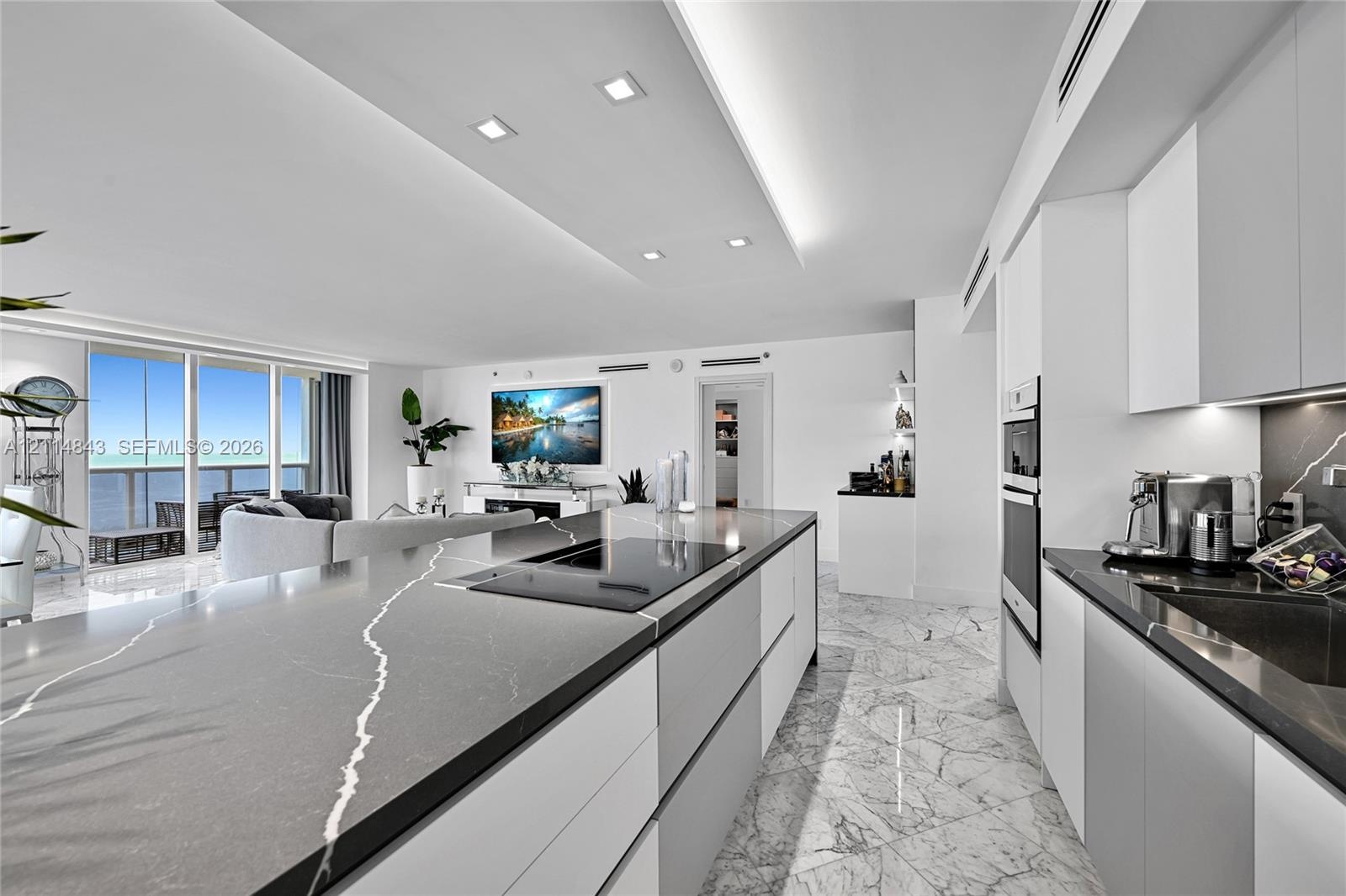 Modern kitchen featuring a large central island with veined stone countertop and integrated cooktop, handle-less cabinetry, built-in double ovens, and