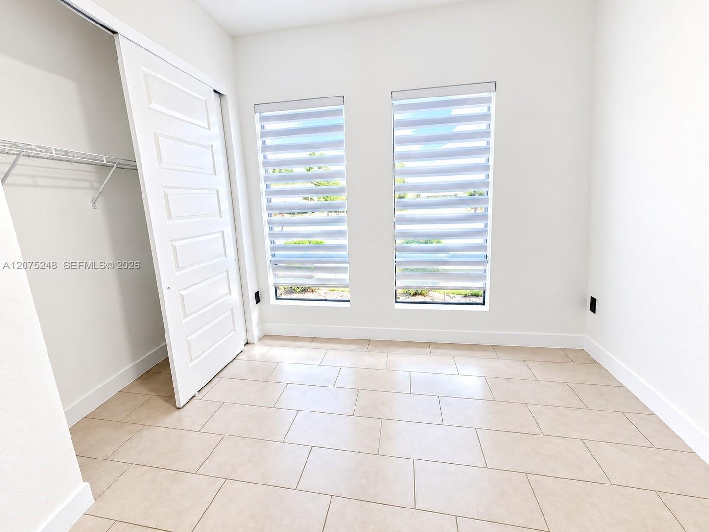 Neutral-toned ceramic tile flooring, dual windows with installed roller shades, and a closet featuring a multi-panel door and wire shelving
