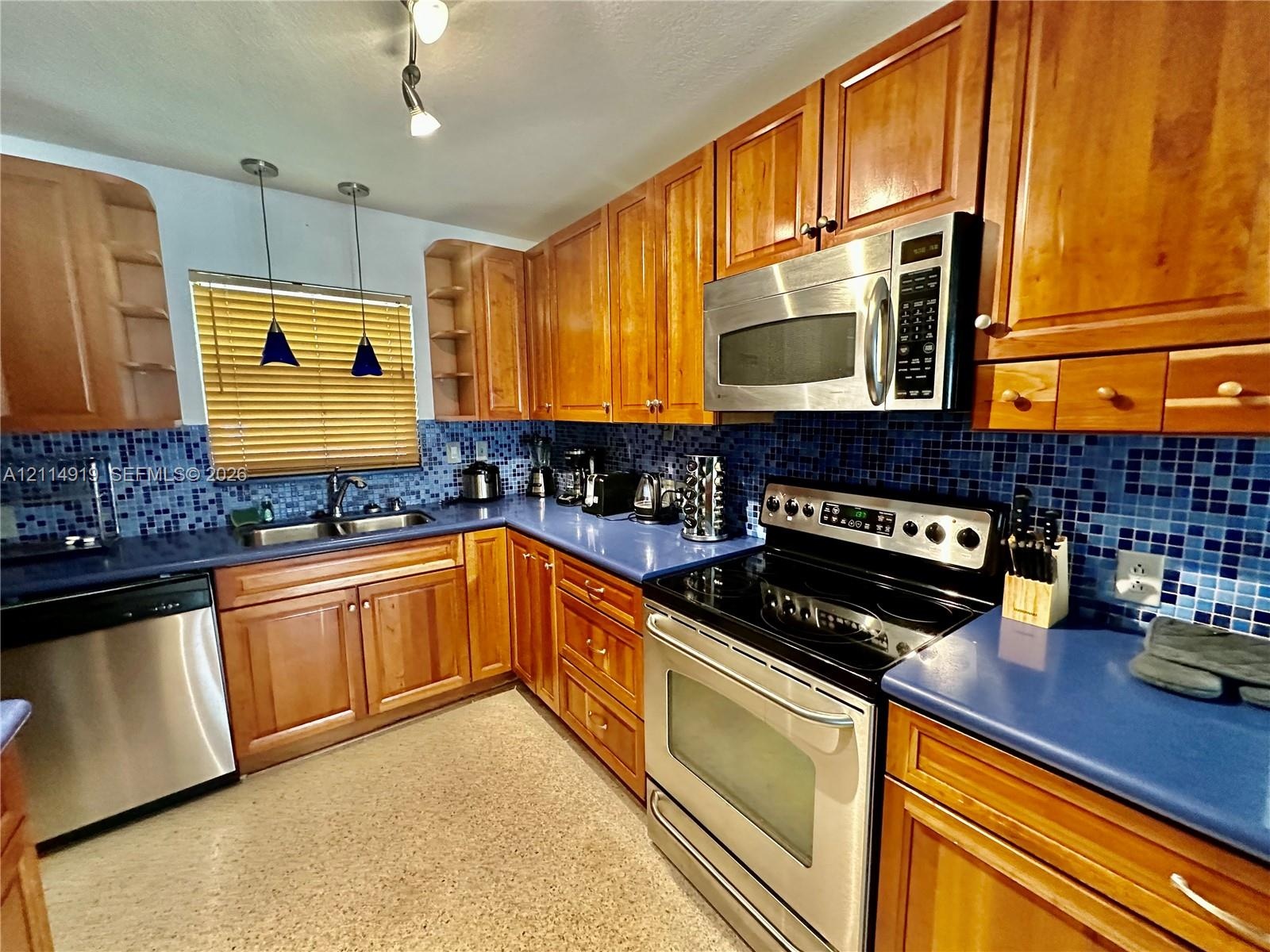 Kitchen featuring wood cabinetry, stainless steel appliances, blue solid-surface countertops, blue mosaic tile backsplash, and dual-basin stainless st