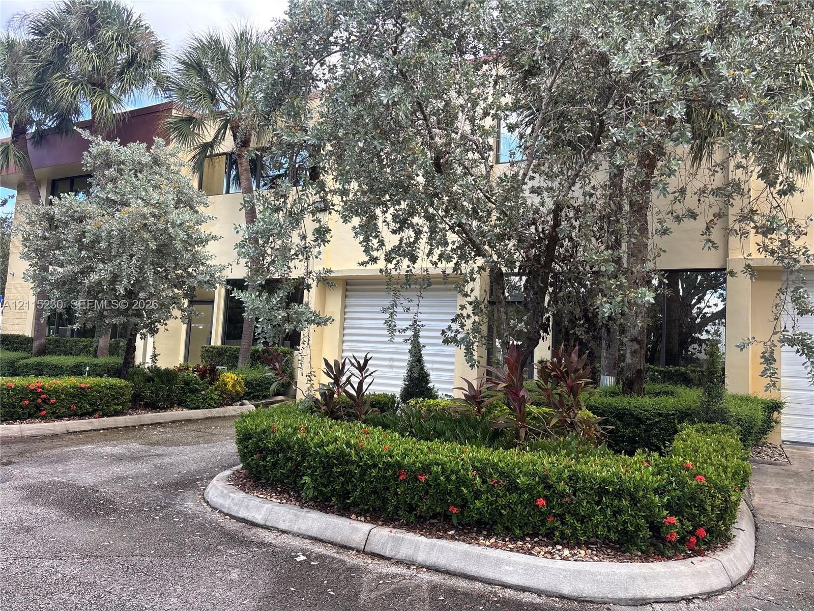 Two-story exterior featuring a light-colored stucco facade, roll-up garage doors, paved driveway, and mature landscaping including manicured shrubs an
