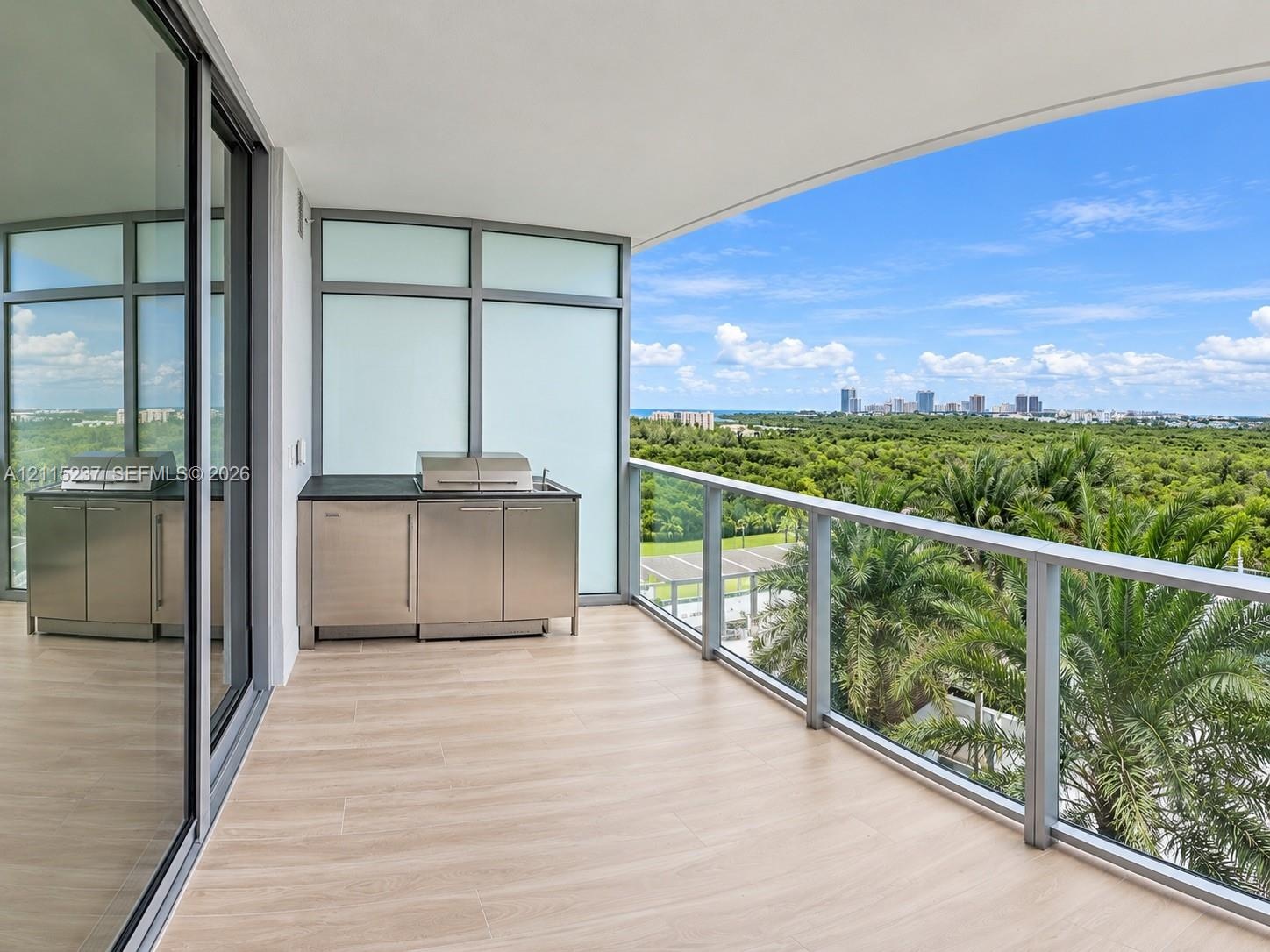 Covered balcony featuring wood-finish flooring, stainless steel outdoor kitchen cabinetry, built-in grill, glass railing, and floor-to-ceiling sliding