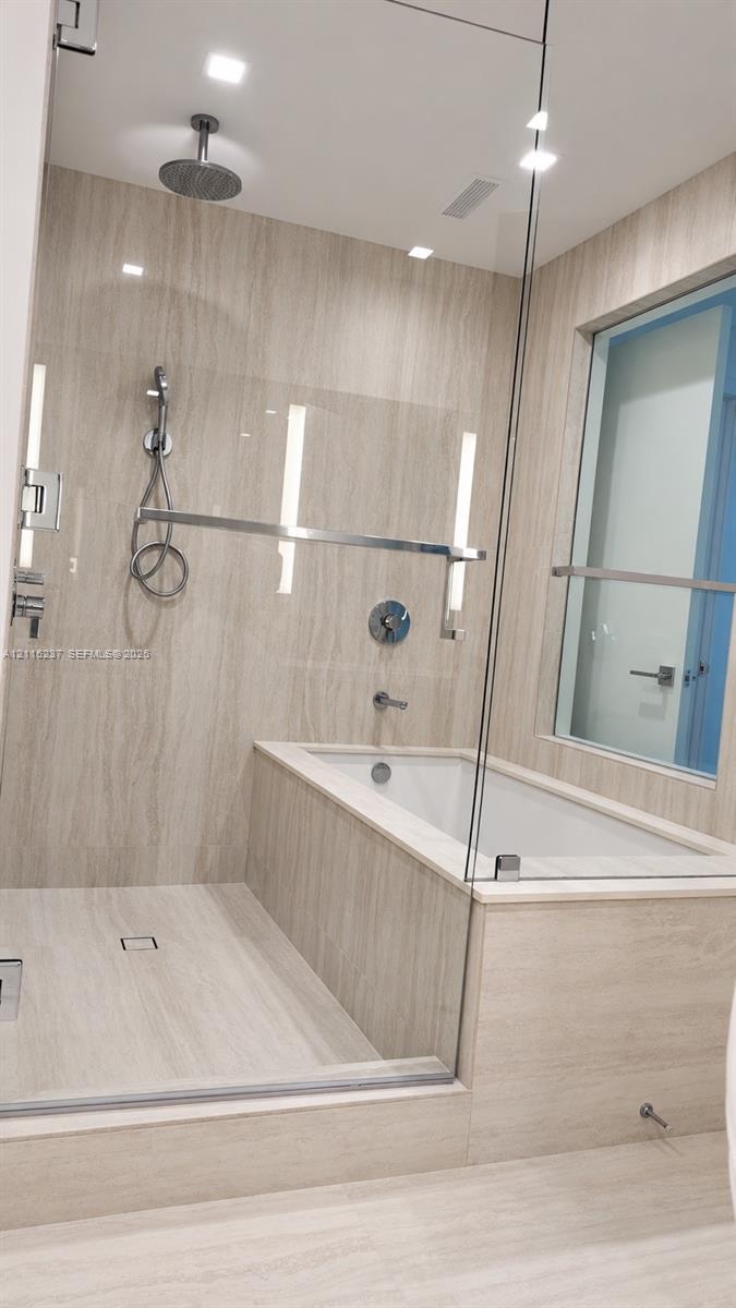 Glass-enclosed wet room featuring a built-in soaking tub, ceiling-mounted rainfall showerhead, handheld shower fixture, and floor-to-ceiling stone-fin