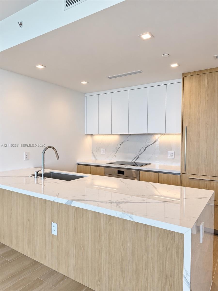 Kitchen featuring a waterfall-edge stone countertop, wood-finish cabinetry, integrated appliances, under-cabinet lighting, and a stone slab backsplash