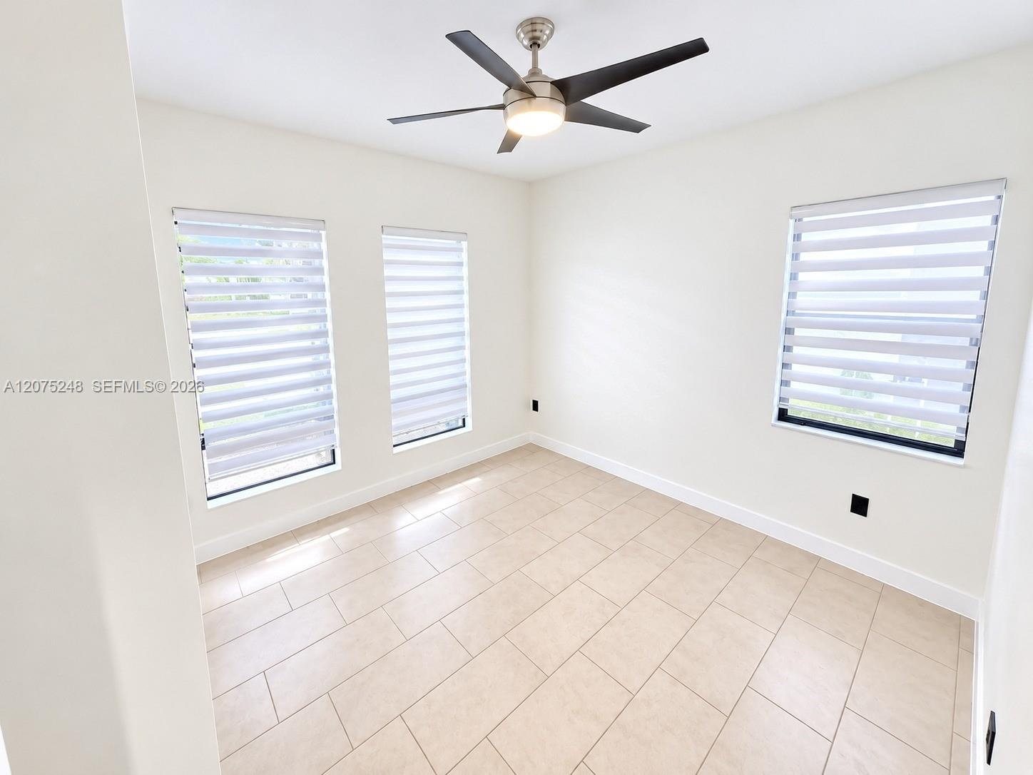 Neutral-toned tile flooring, three windows with adjustable roller shades, and a ceiling-mounted light fixture with a multi-blade fan