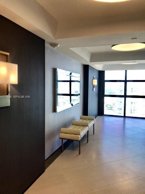 Large-format tile flooring, recessed tray ceiling, floor-to-ceiling windows, dark wood-finish wall paneling, and wall-mounted light fixtures