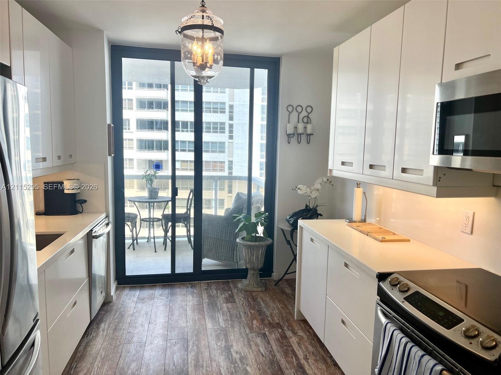 Galley kitchen featuring high-gloss white cabinetry, solid-surface countertops, wood-finish flooring, stainless steel appliances, and a glass-paneled 