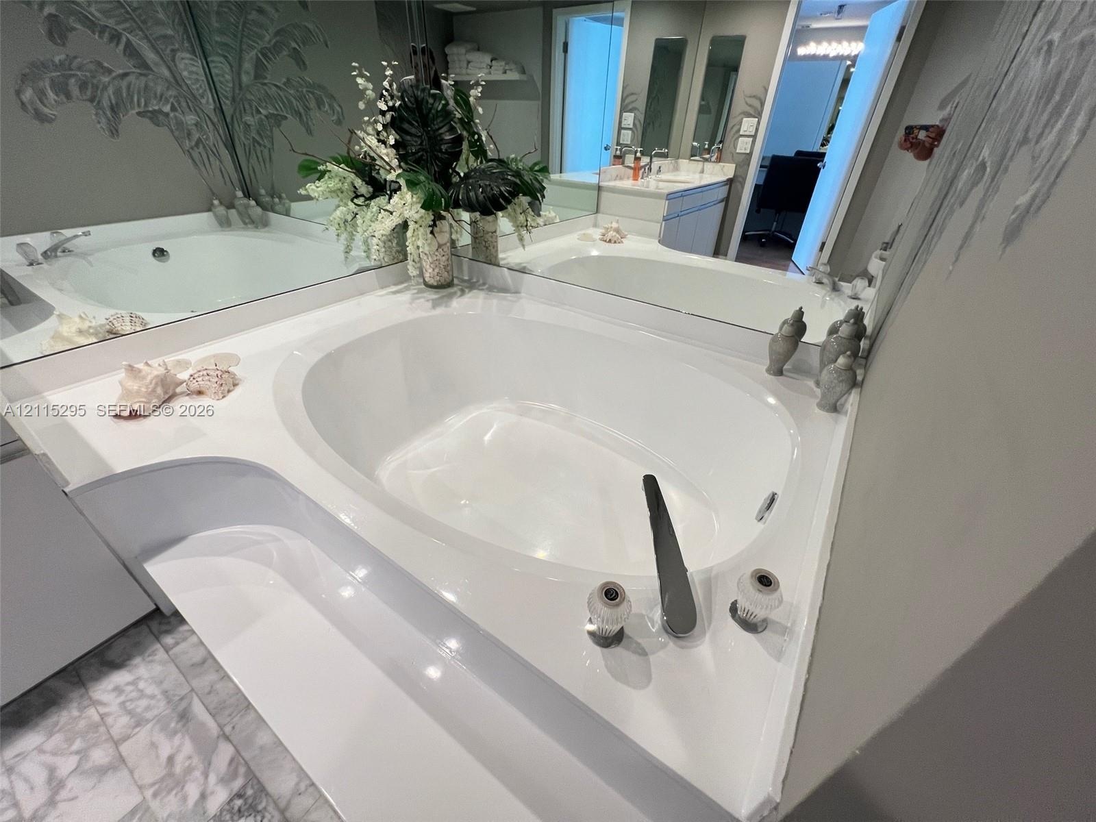 Oval soaking tub with a deck-mounted faucet and hand shower, set into a solid-surface surround, complemented by a large wall-to-wall mirror and natura