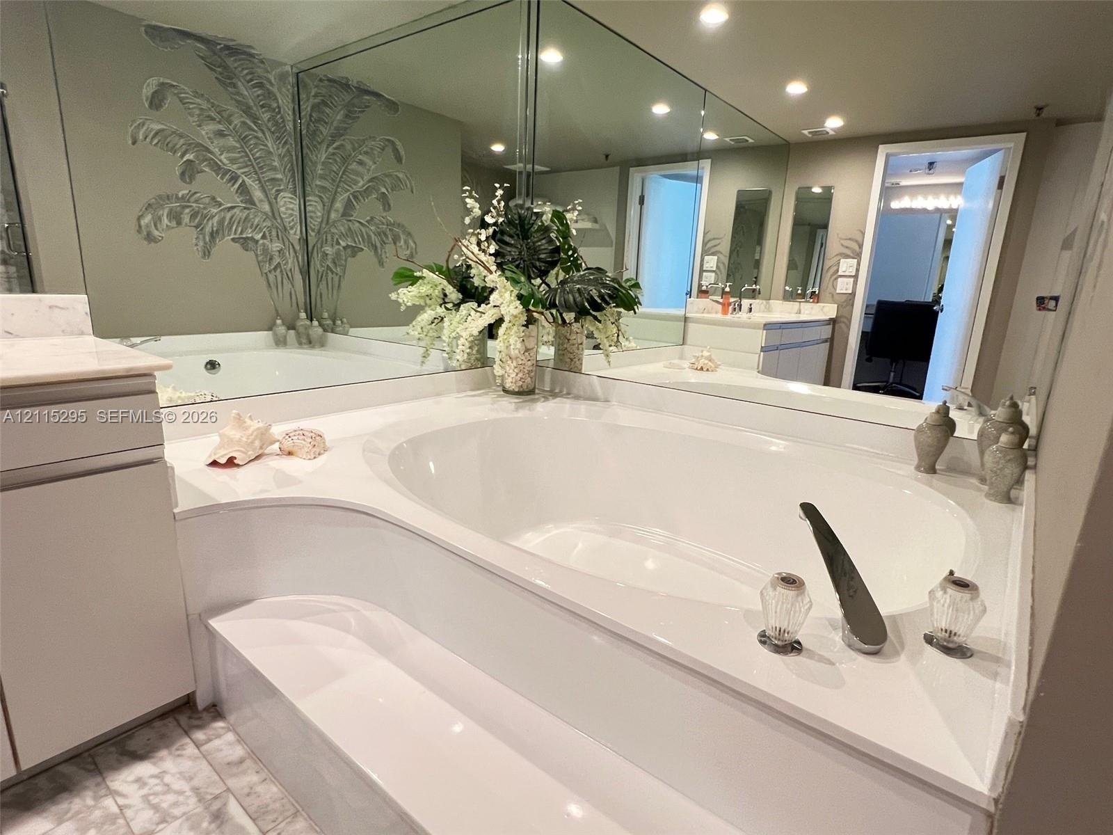 Oval soaking tub with integrated deck, large-format wall-to-wall mirror, built-in cabinetry, and tile flooring