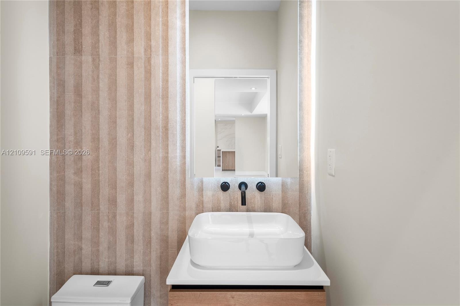 Half Bath off living room. Wall-mounted matte black faucet, white rectangular vessel sink, white countertop, wood-finish vanity, and vertical textured