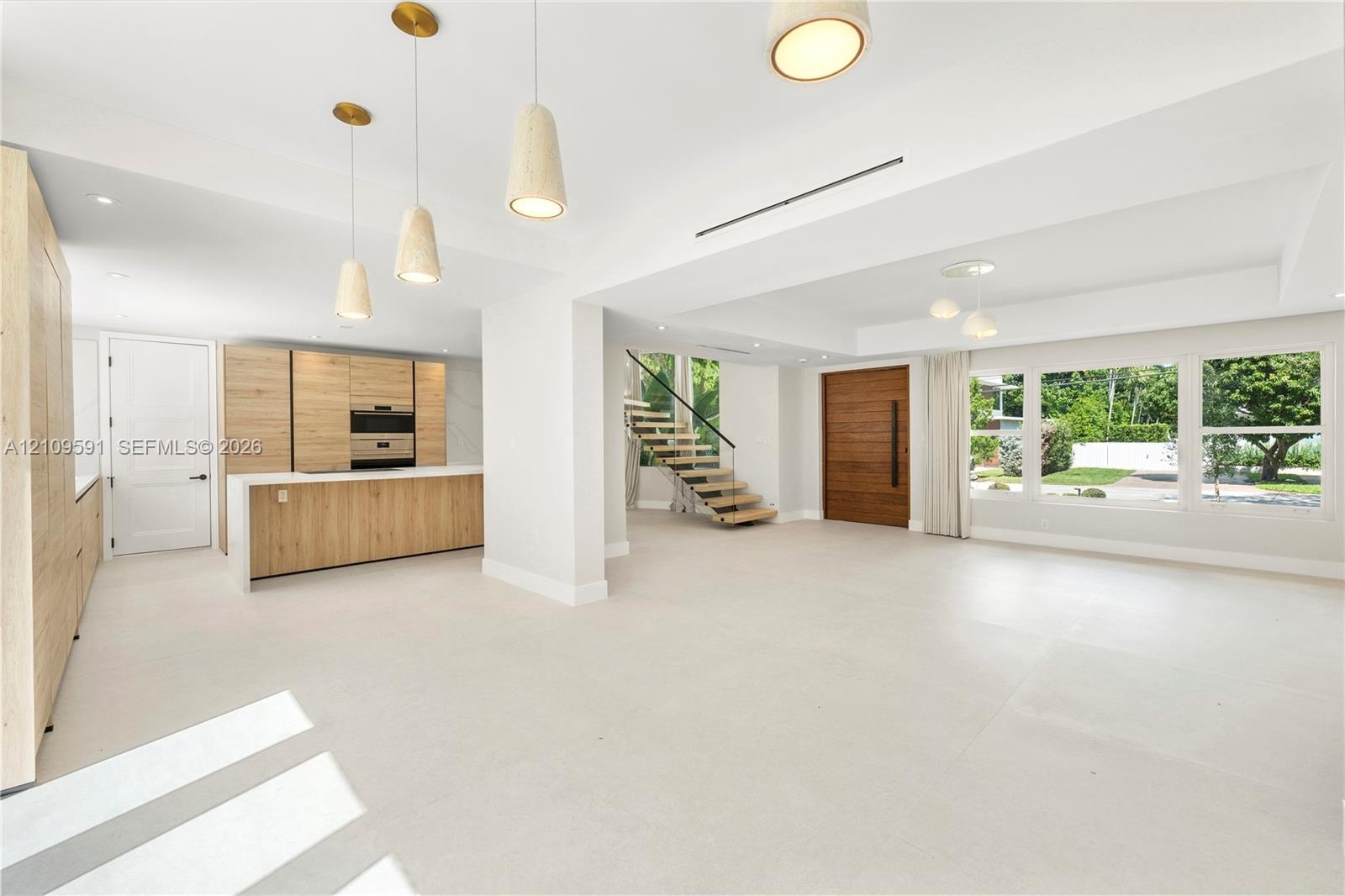 Open-concept interior featuring wood-finish cabinetry, a central kitchen island, large-format tile flooring, a floating staircase with glass railing, 